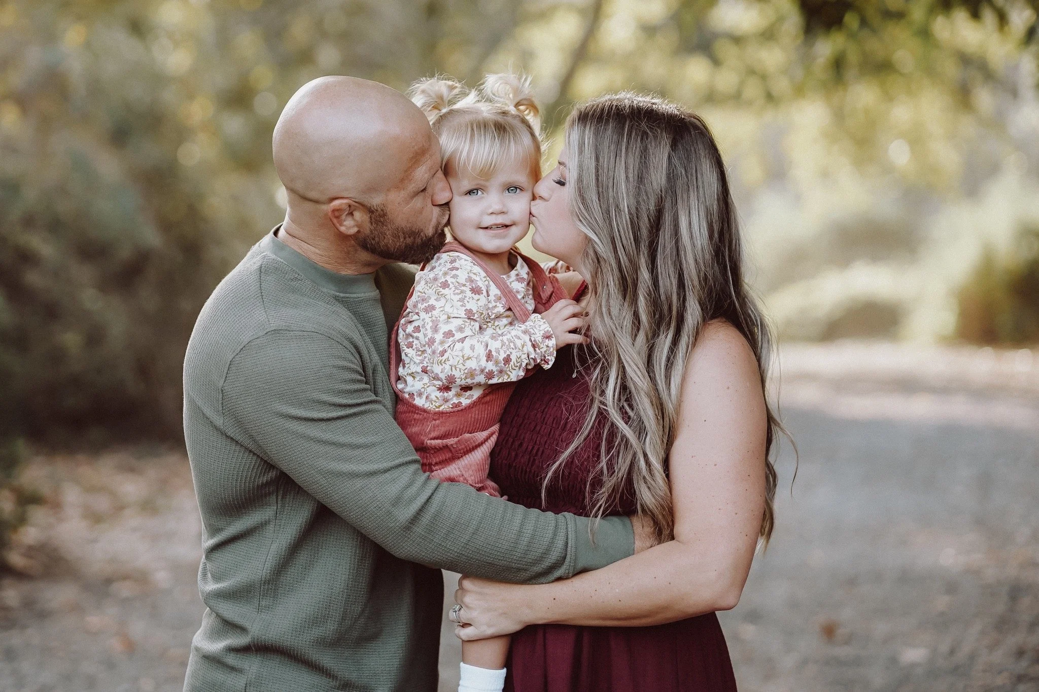 Mom and dad holding toddler while kissing her on the cheek