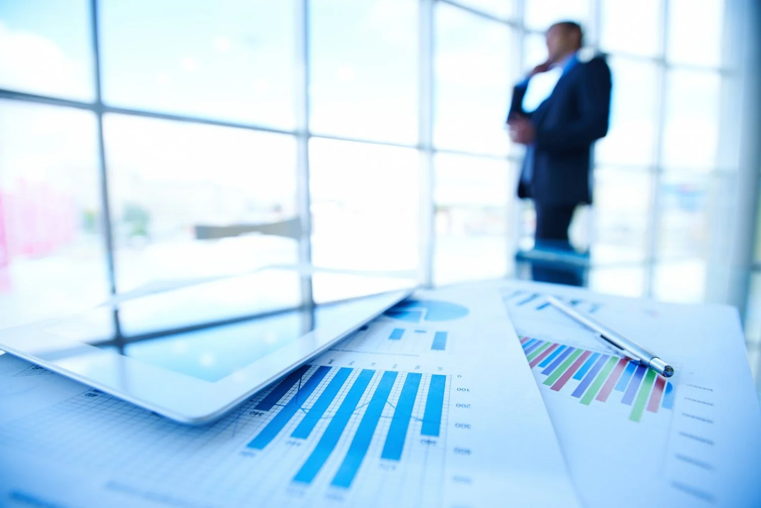 Financial charts and reports spread on a desk with a tablet and pen in the foreground, while a blurred professional stands near large office windows in the background.