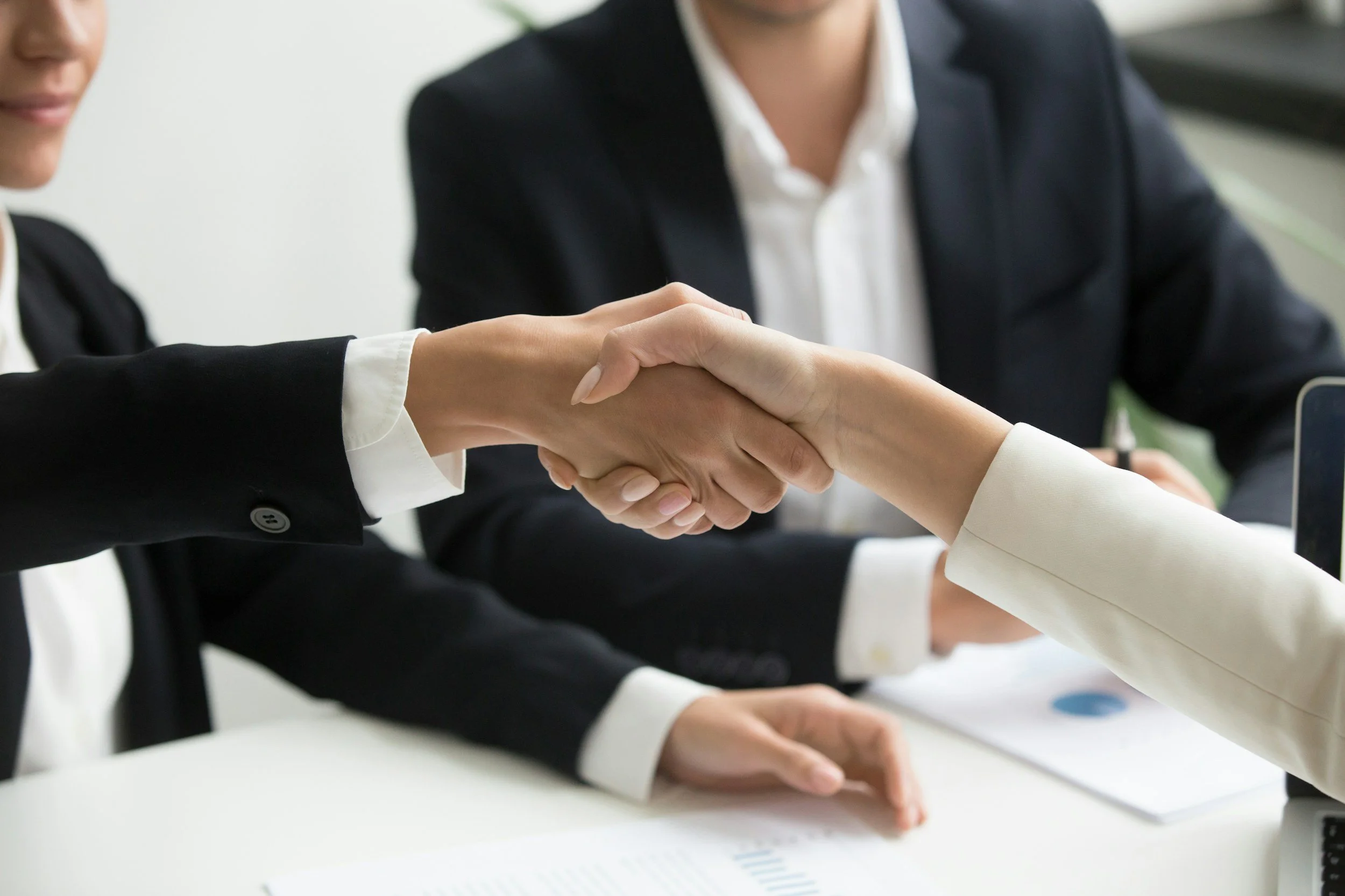 Two women in business suits shaking hands during a meeting, with another person sitting nearby.