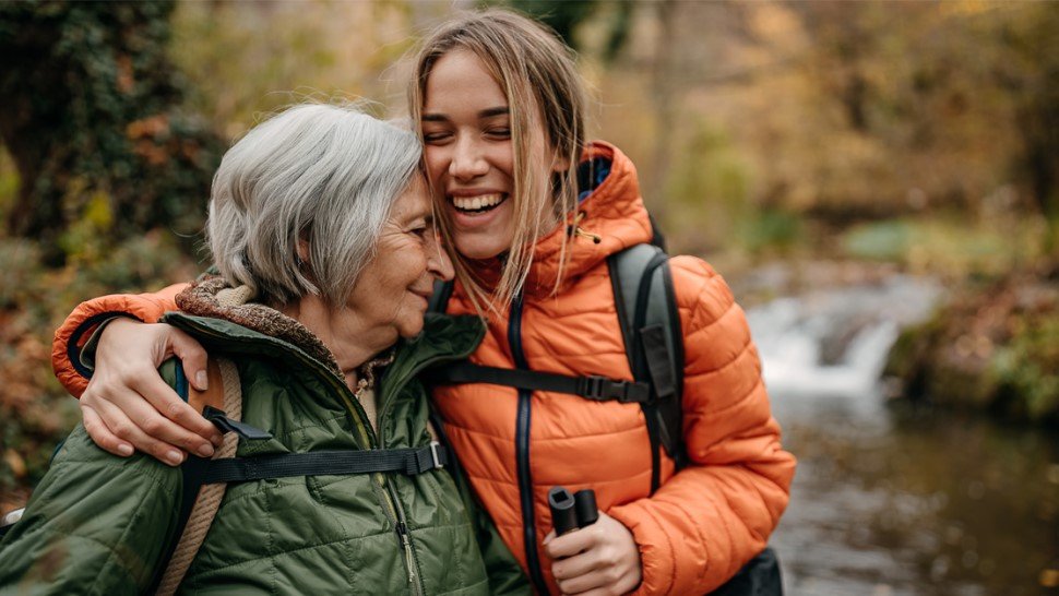 Smiling older woman and young woman embrace on an autumn hike near a small waterfall.
