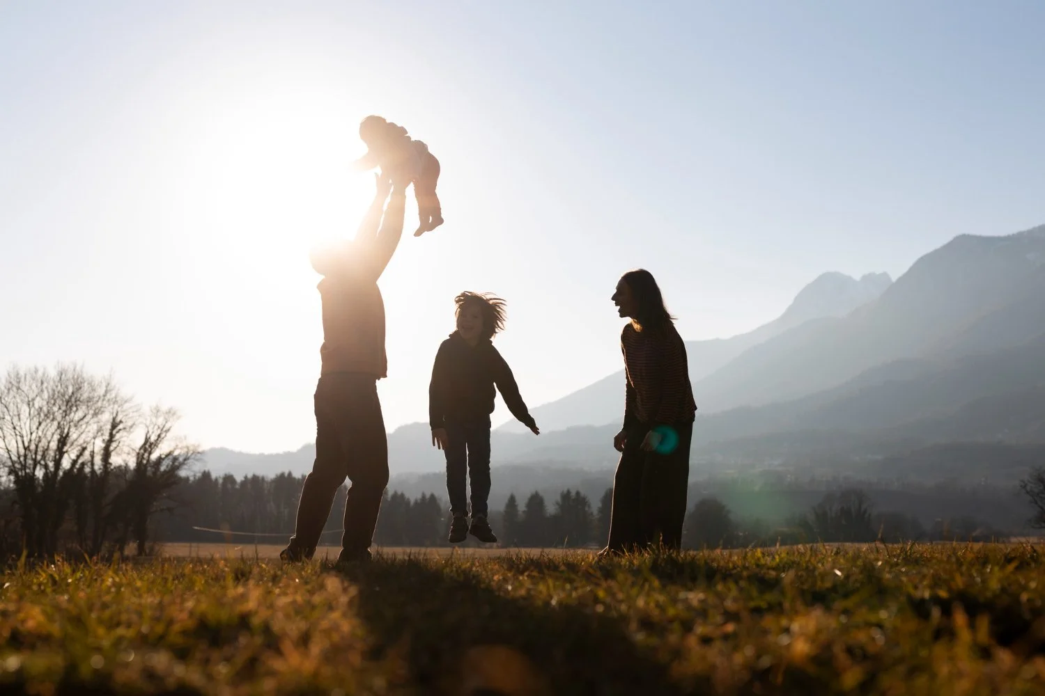 A family plays together outdoors at sunset, with a parent lifting a baby into the air while another adult and a child jump and laugh in an open field with mountains in the background.