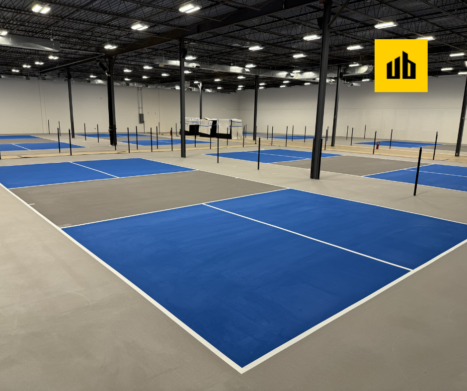 Empty indoor pickleball courts with blue and gray surfaces, black net posts, and a yellow logo in the top right corner.