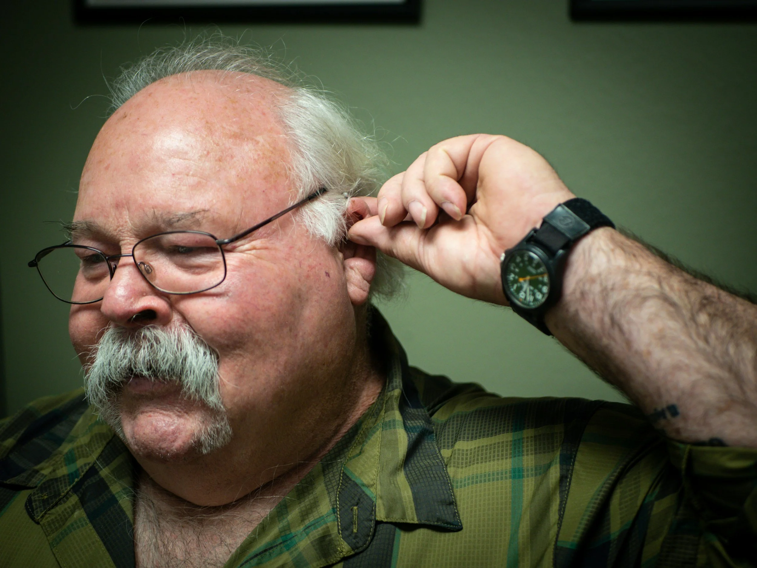 Man in a green shirt in front of a green background putting in a behind the ear hearing aide