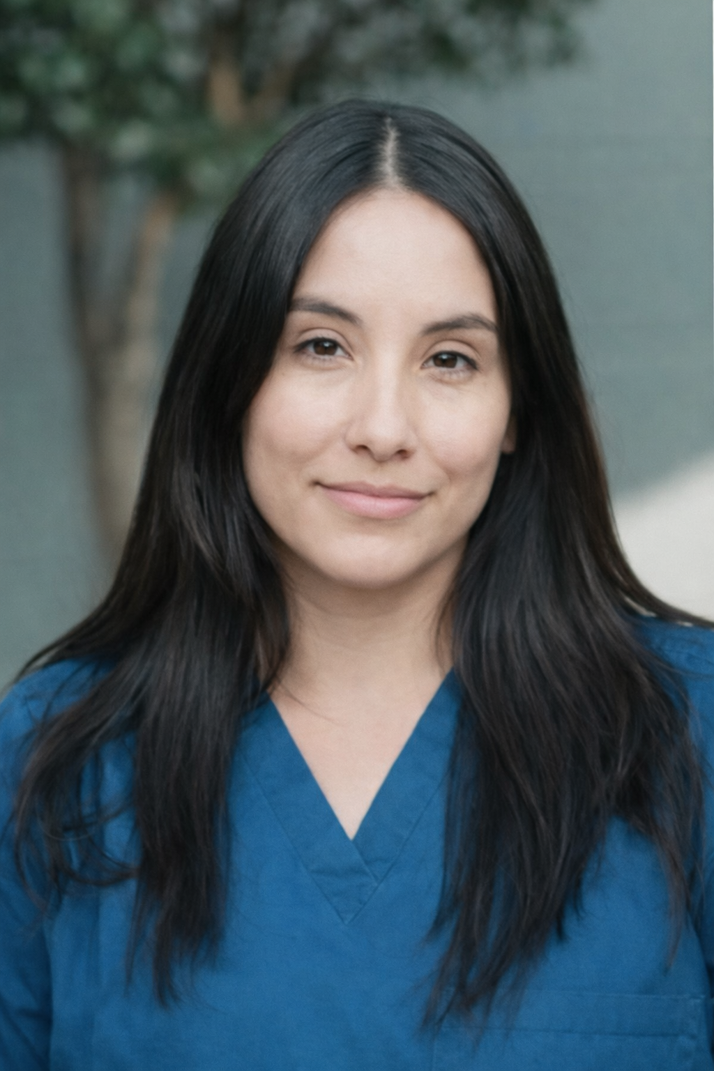 A woman smiling outdoors wearing blue medical scrubs with the word "RESTED" embroidered on it, with trees and foliage in the background.