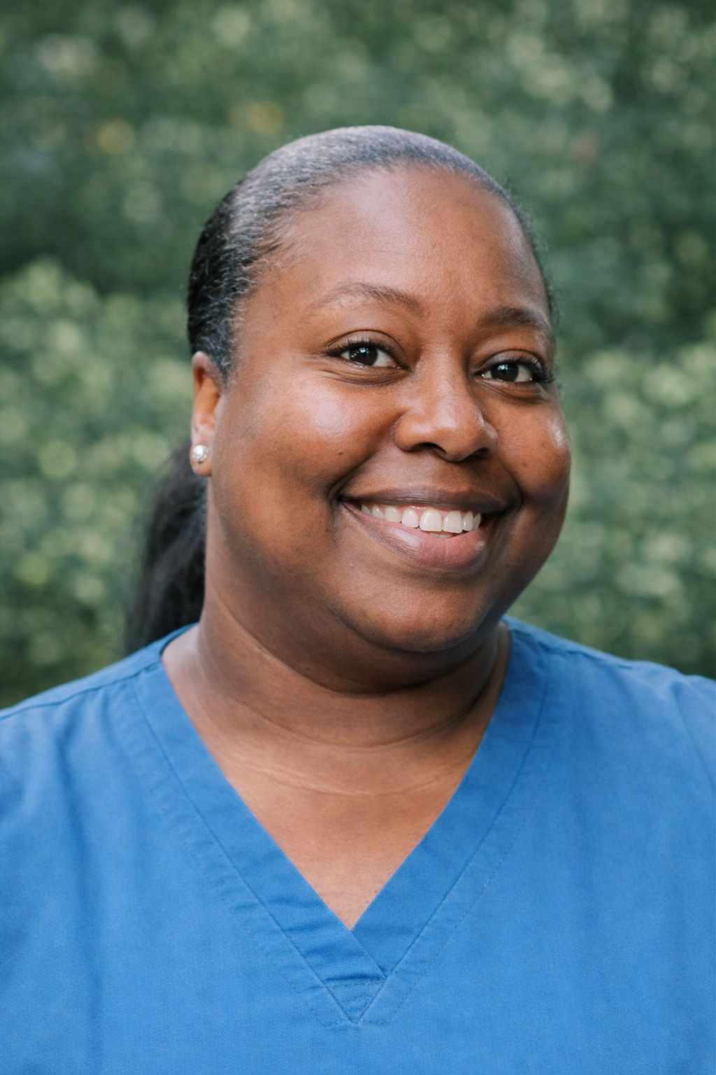A woman wearing blue medical scrubs, standing outdoors with greenery and trees in the background, smiling at the camera.