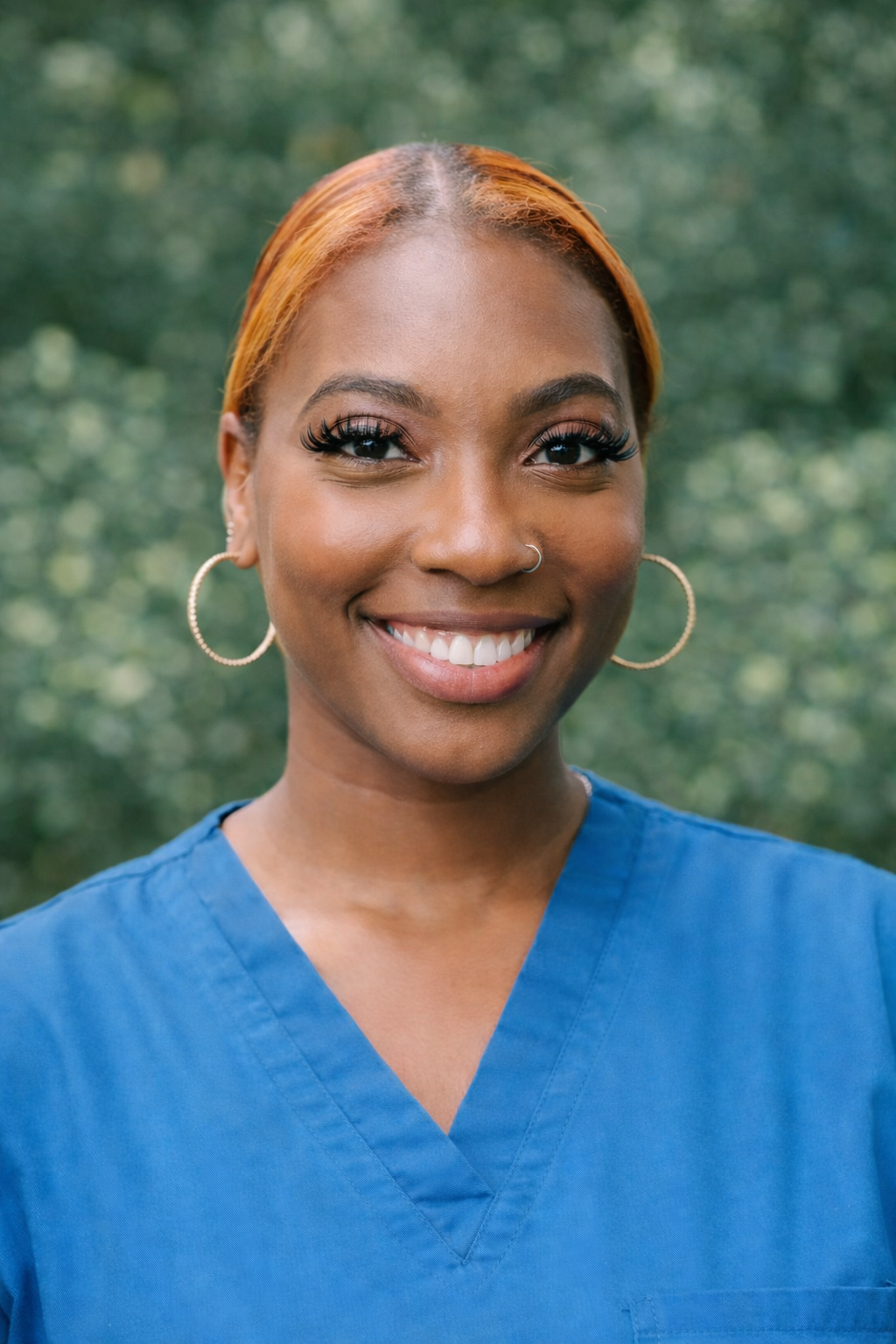 A young African American woman with short hair, gold hoop earrings, and a nose ring, wearing navy medical scrubs, smiling outdoors with blurred greenery in the background.
