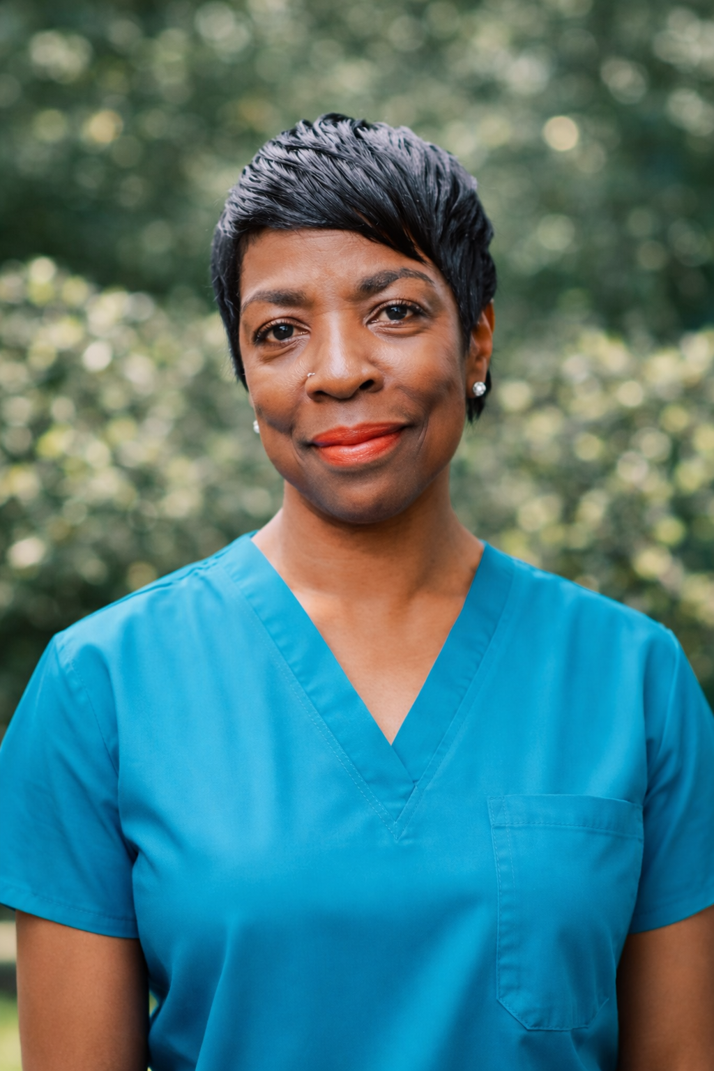 A smiling woman in blue scrubs standing outdoors with green trees in the background.