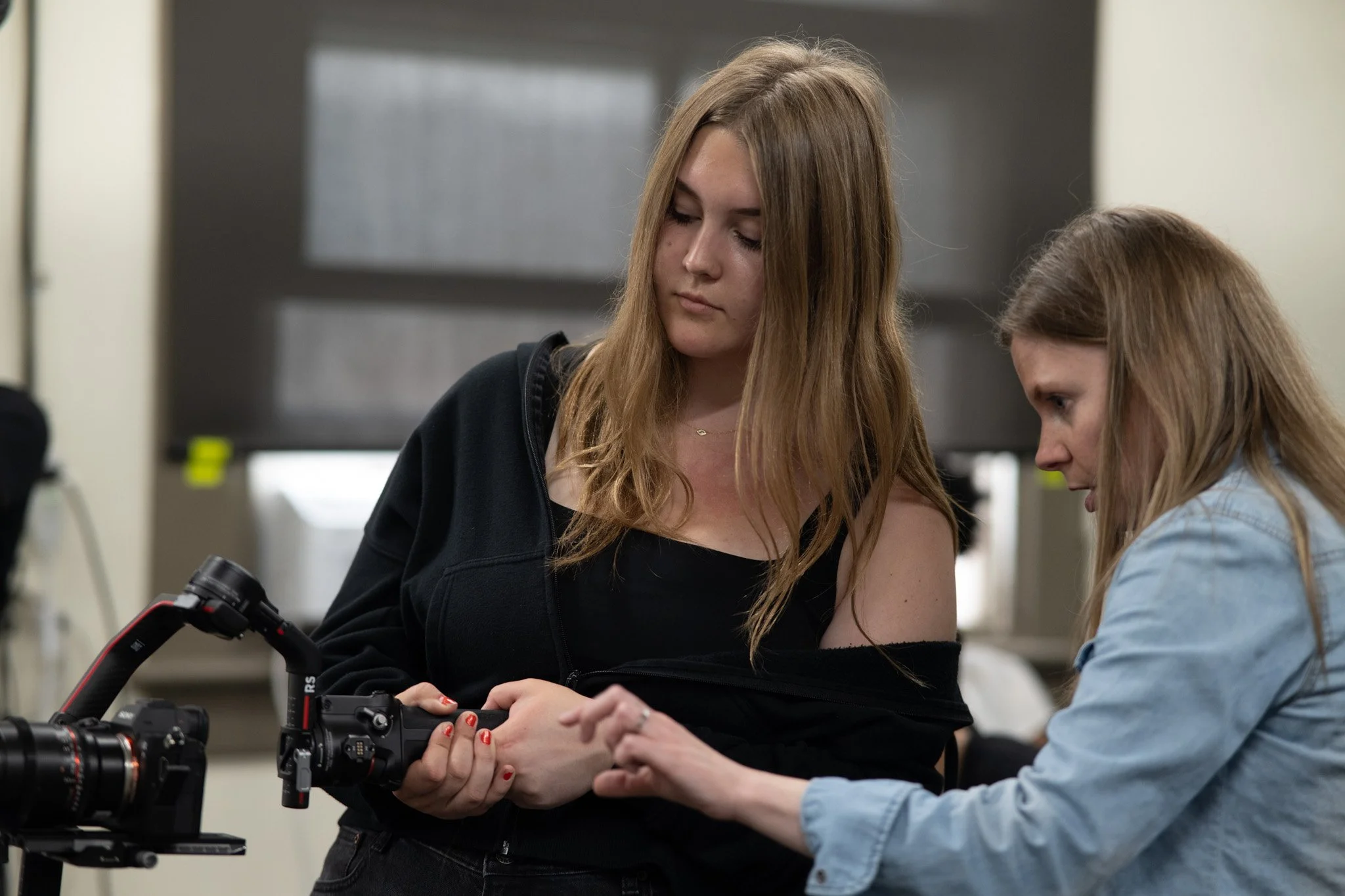 Two women are working together on a camera in an indoor setting. One woman has long, light brown hair and is wearing a black jacket, while the other has long, blonde hair and is wearing a light blue shirt. They are focused on the camera equipment on the table in front of them.
