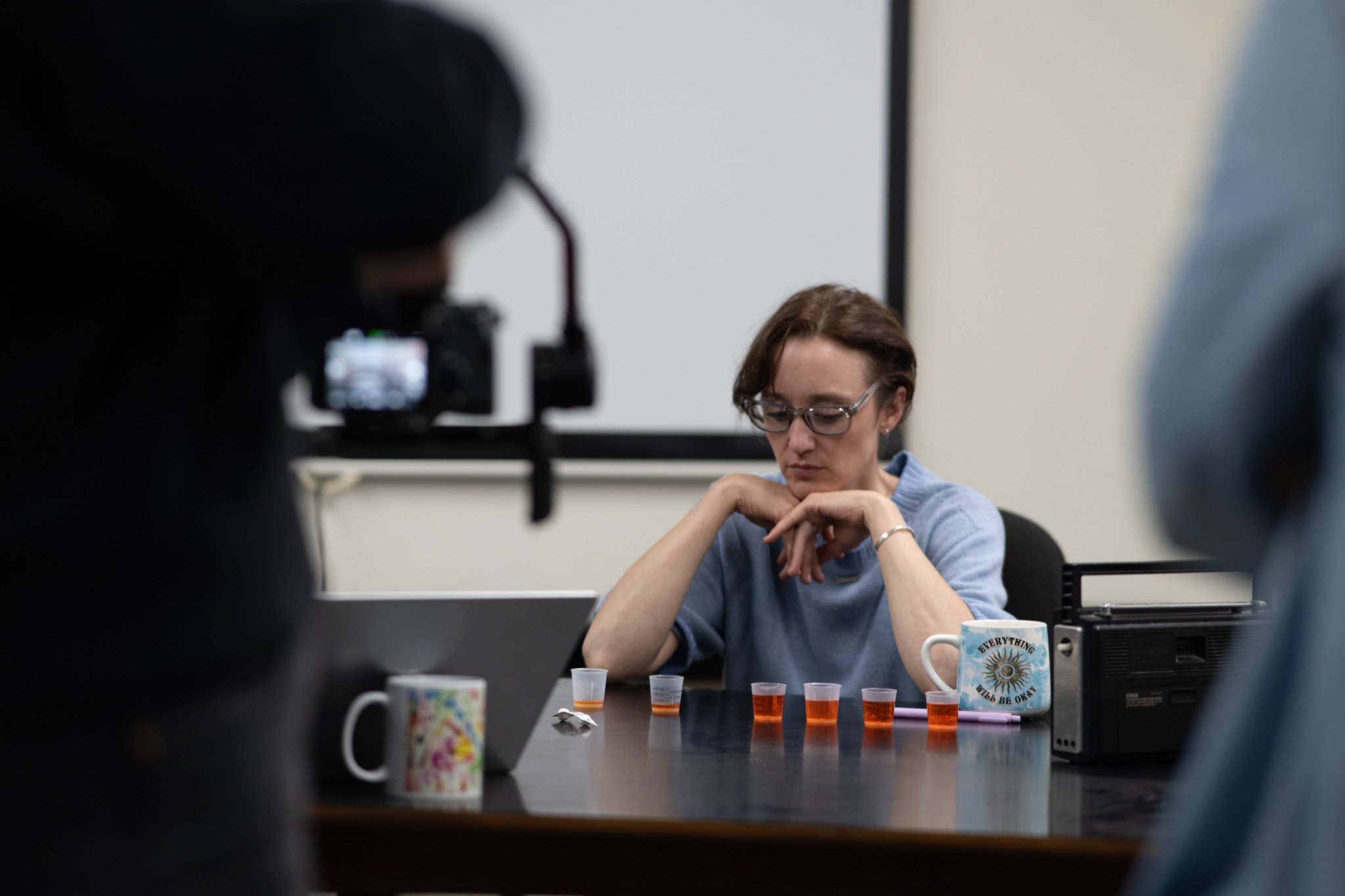 A woman with short brown hair and glasses sits at a table, looking down with her hands clasped, surrounded by cups and small containers. A camera on a tripod captures her from the left side of the image.