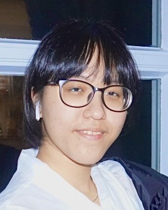 A person with short black hair, glasses, and wireless earbuds, smiling at the camera indoors near a window.