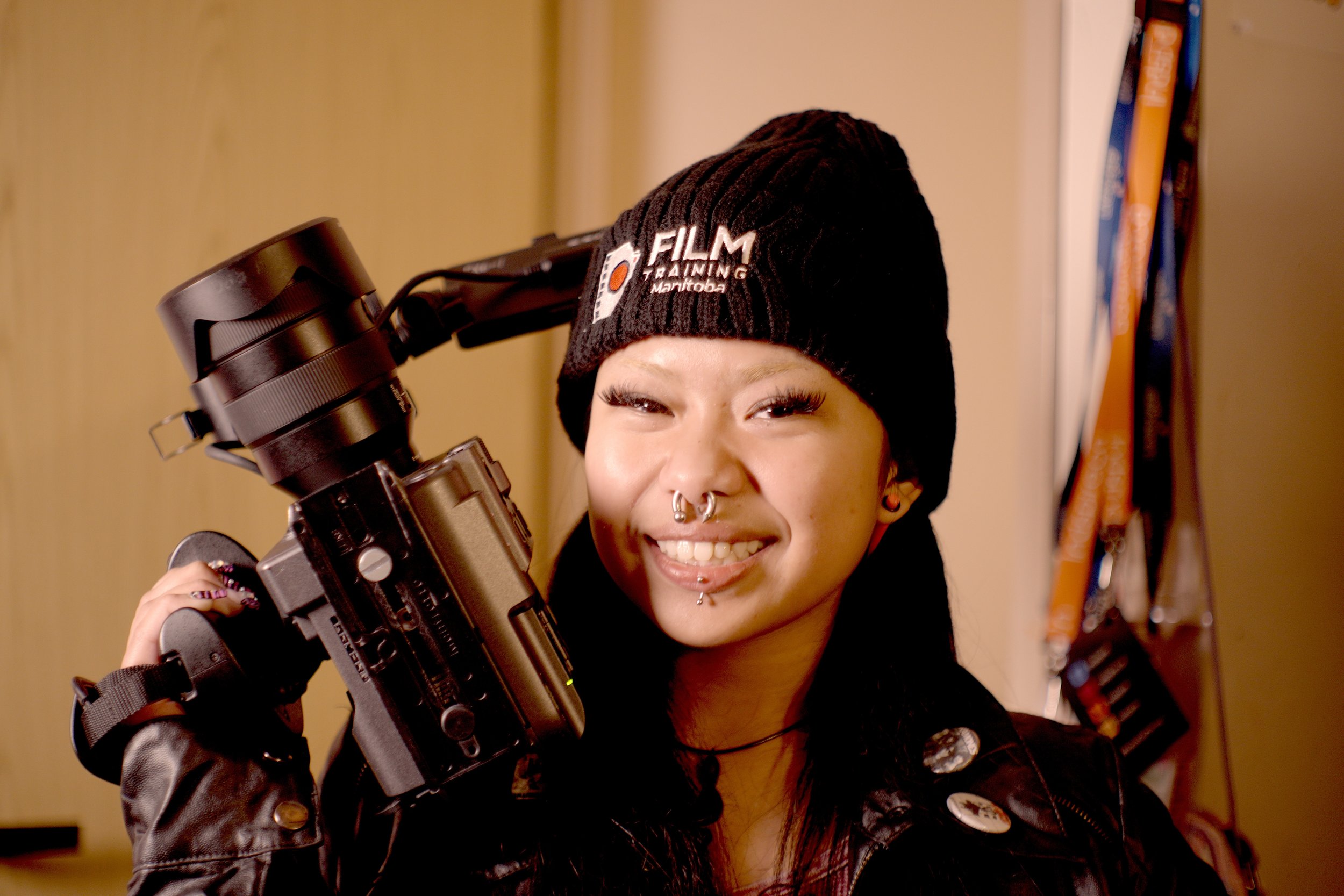 A woman with multiple facial piercings smiling while holding a camera, wearing a black beanie with 'Film Training Manitoba' embroidered on it, and a black leather jacket.