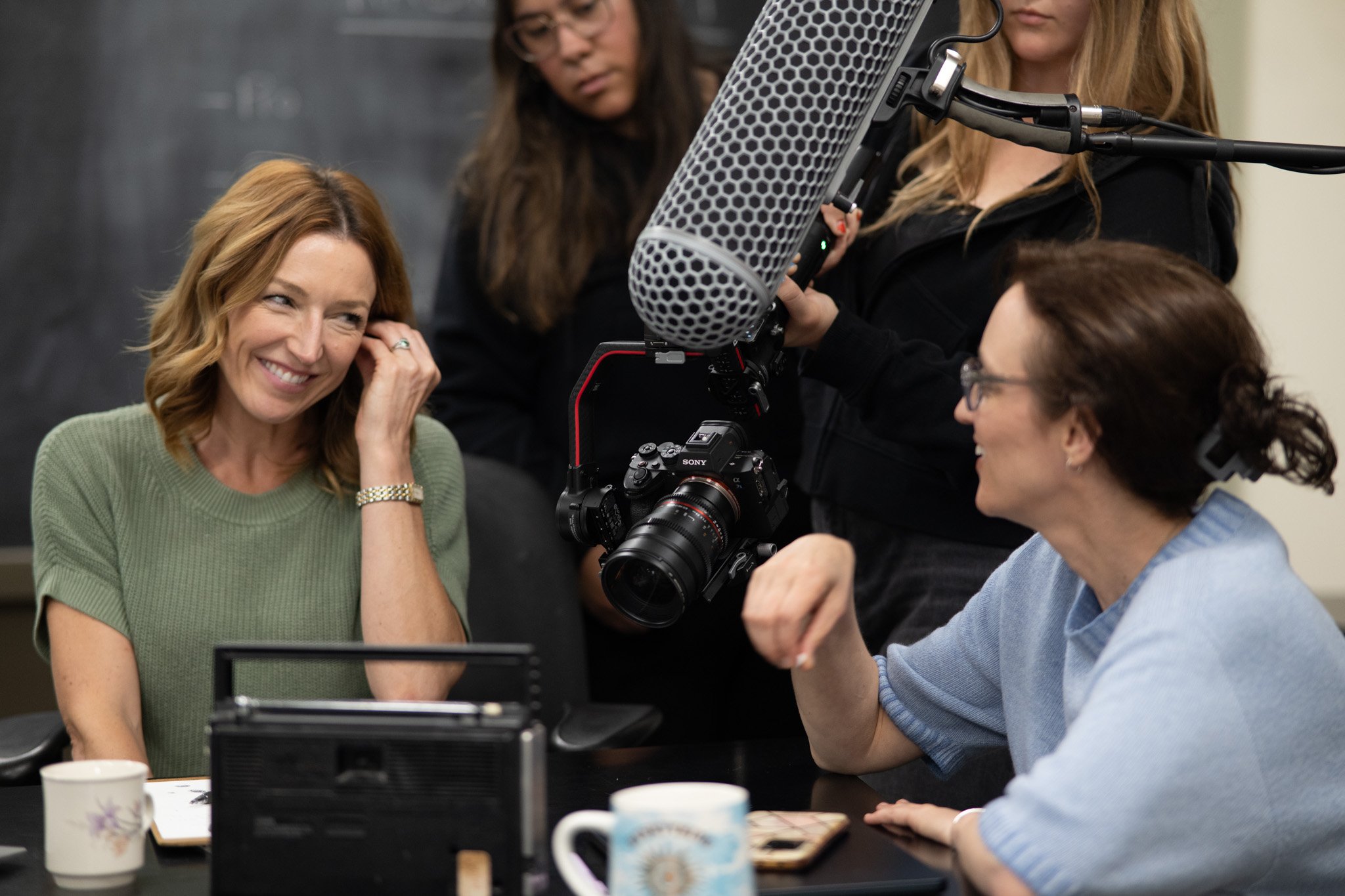 A woman with red hair smiling and adjusting her earring while seated at a table with three mugs, with a film crew working behind her, including two women operating a camera and microphone.