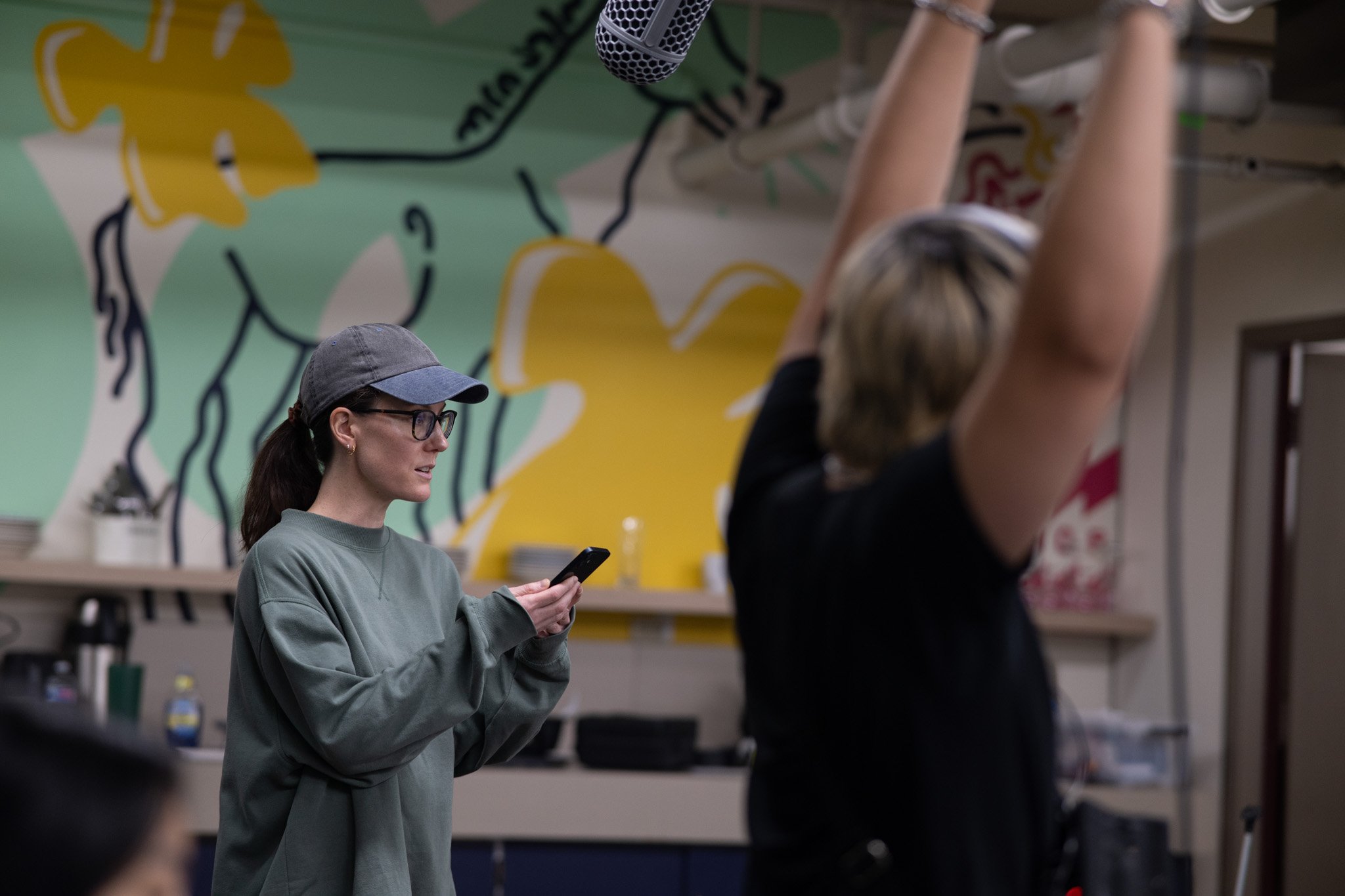 A woman wearing glasses, a gray hoodie, and a gray cap, looks at her smartphone while standing in what appears to be a creative workspace or classroom. In the background, another person with blonde hair tied back is raising their arms, and the wall has colorful, abstract graffiti-style artwork.