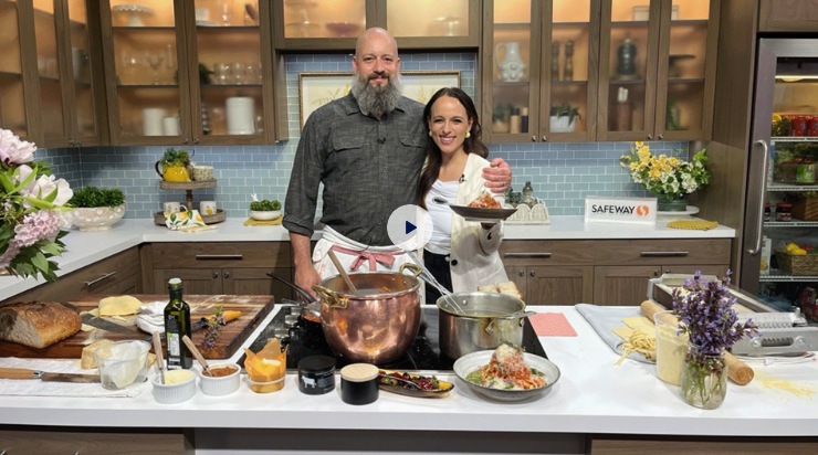 A man and woman standing in a kitchen, smiling at the camera as they prepare a meal. The kitchen is well-lit with wooden cabinets and various cooking ingredients and utensils on the counter.