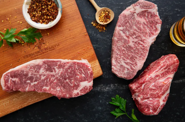 Three raw marbled steaks on a dark surface, with a wooden cutting board, a small bowl of seasoning, a spoon, a sprig of parsley, and a small bottle of seasoning or oil nearby.