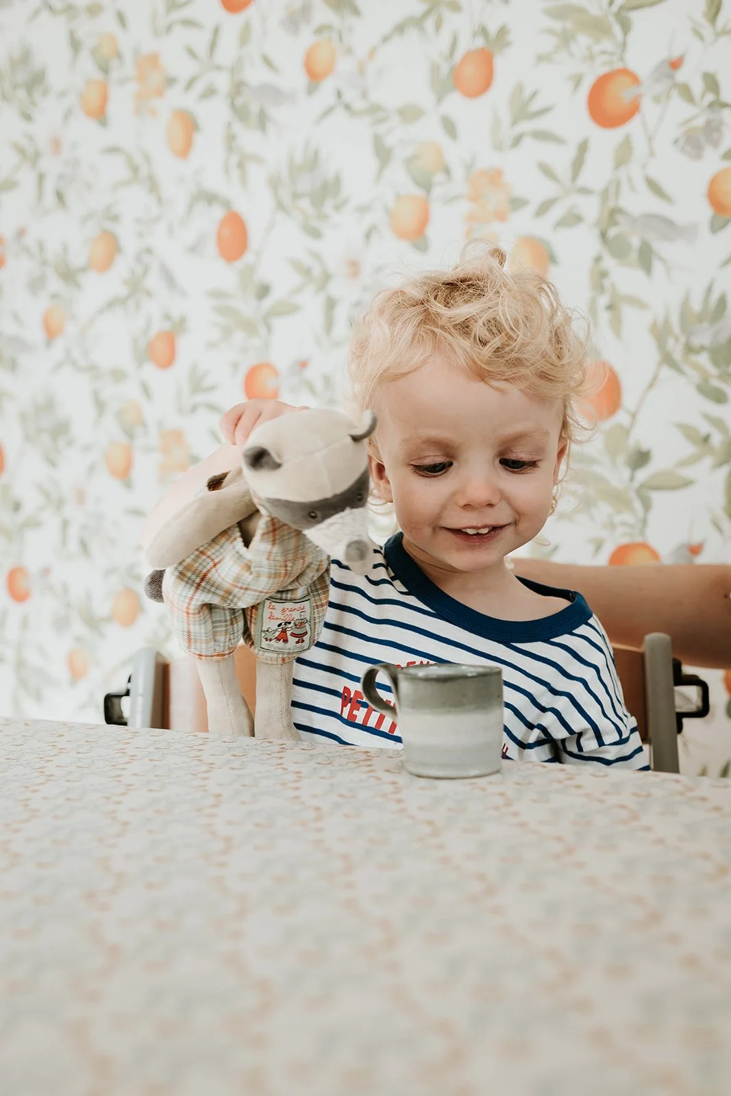A young boy with curly blond hair smiling and playing with a stuffed animal of a raccoon at a table with a mug in front of him, against a background of orange and green fruit-patterned wallpaper.