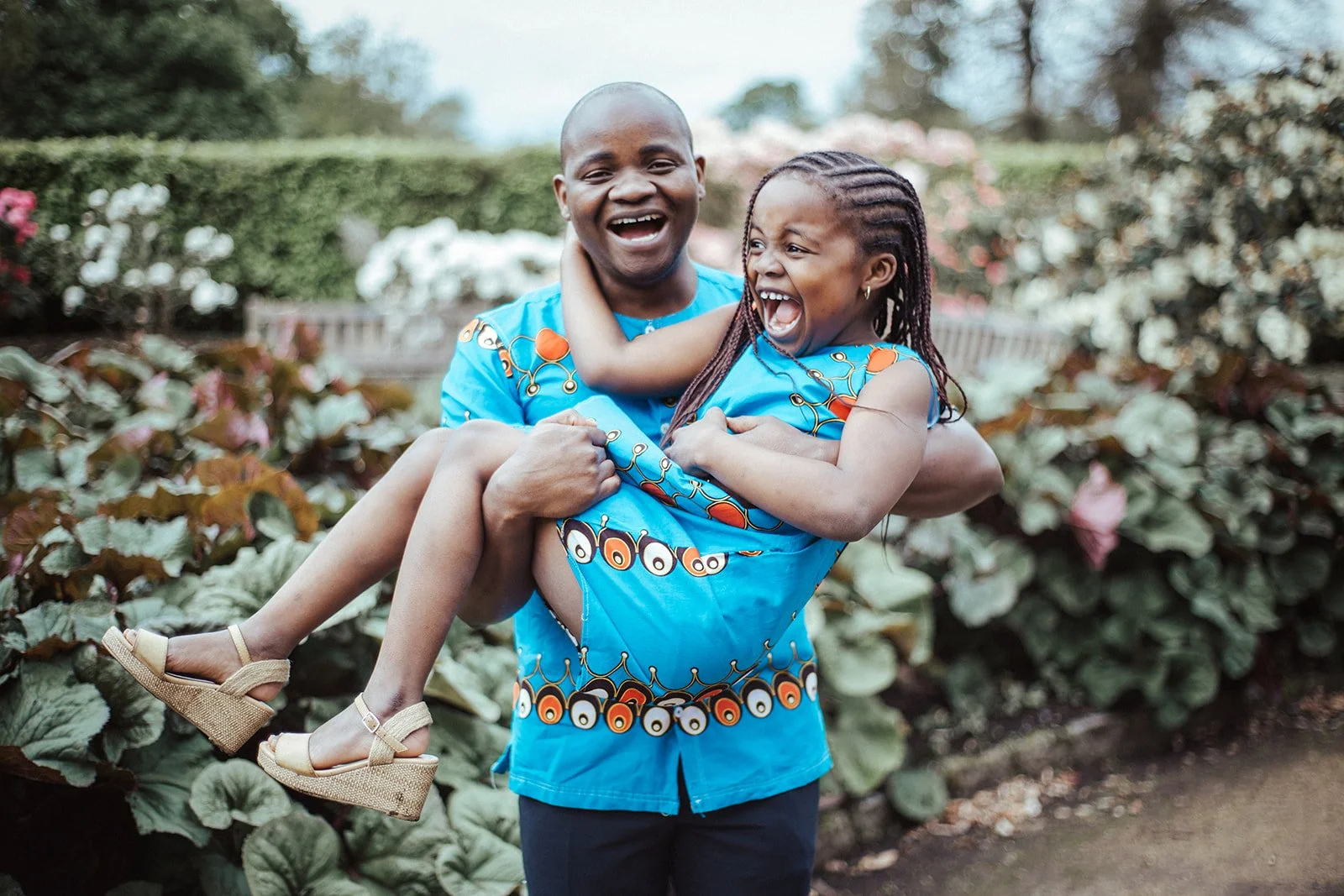 A man holding a young girl in his arms, both wearing matching bright blue traditional African attire, smiling and laughing outdoors in front of bushes with flowers.