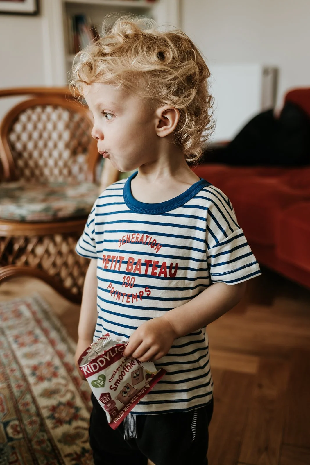 A young boy with curly blonde hair, wearing a striped T-shirt, standing indoors with a partially eaten snack in his hand, looking to the side.