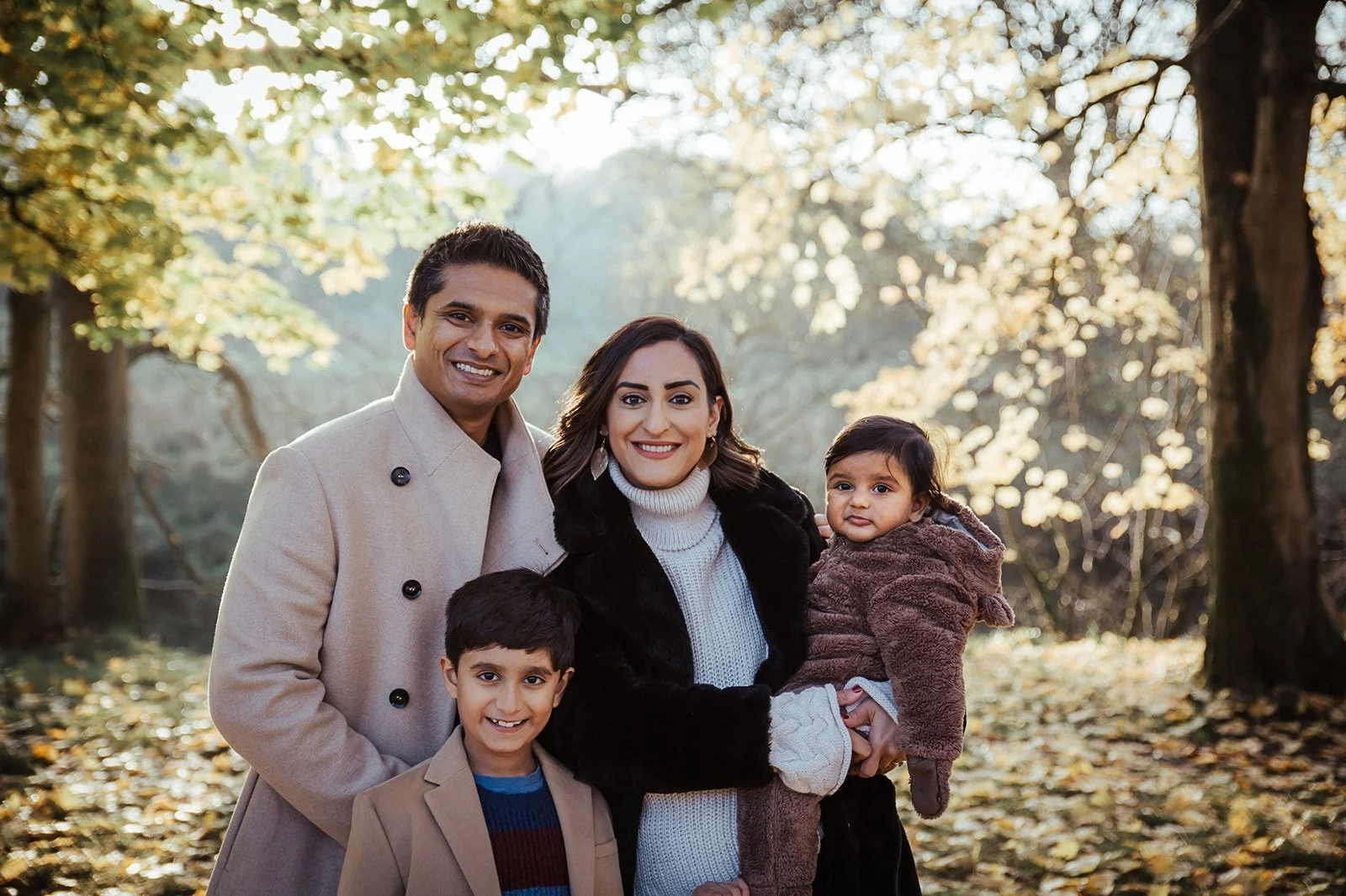 A happy family of four standing outdoors in a park during autumn, with trees and fallen leaves surrounding them. The father, mother, young boy, and toddler girl are smiling at the camera.