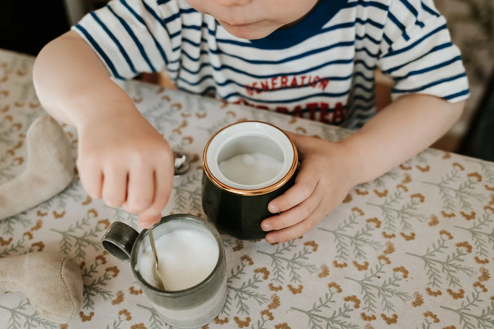 Child in a striped shirt pouring milk from a glass bottle into a jar on a table with a patterned tablecloth.