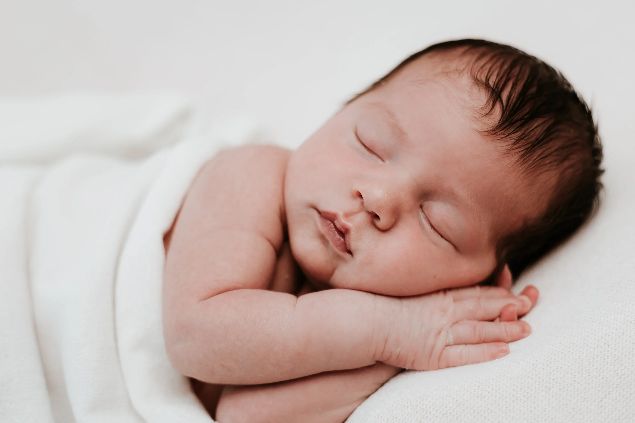 A sleeping newborn baby with dark hair rests on its side, curled up with arms folded close to the face, on a soft white surface.