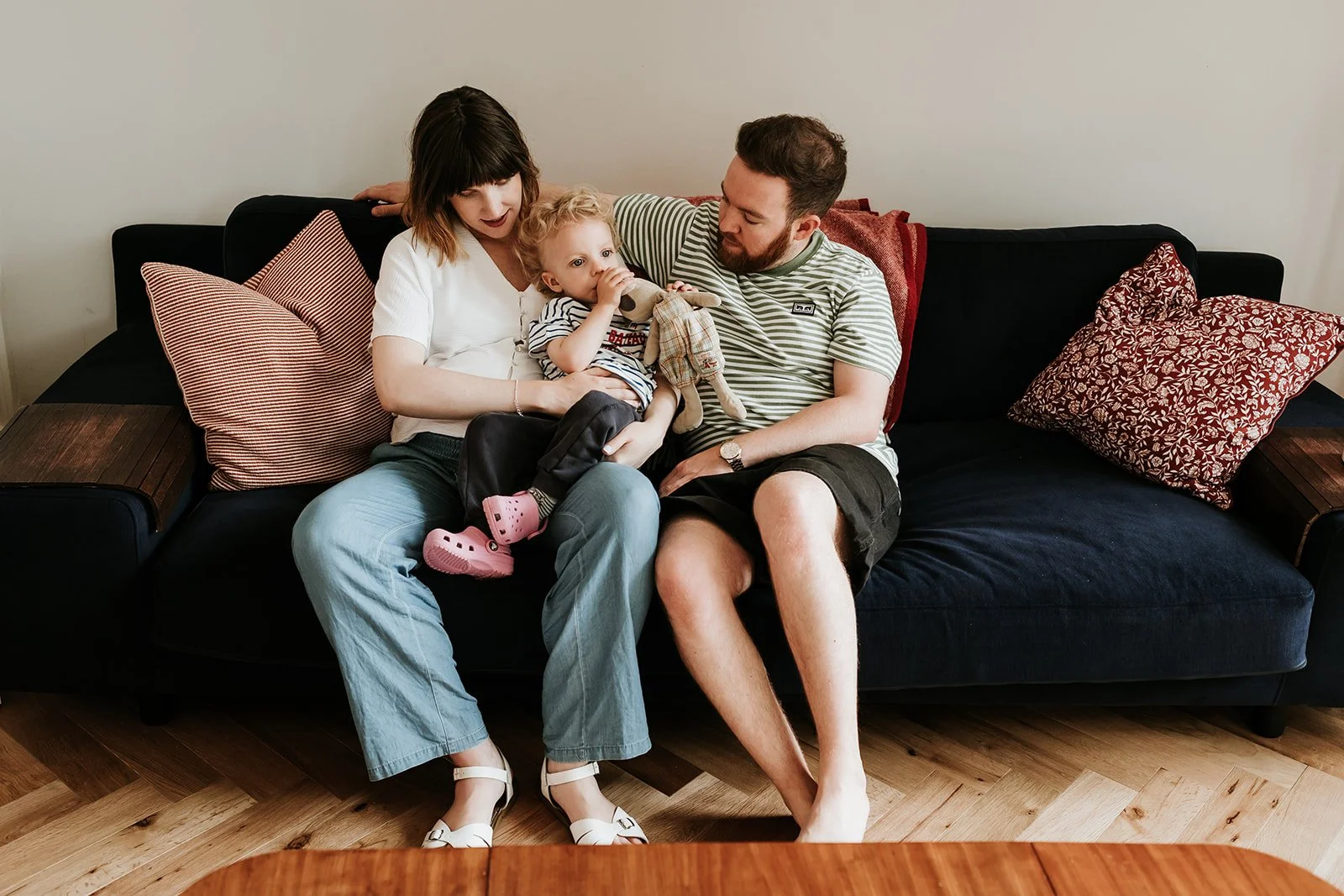 A family of three sitting on a dark-colored sofa with patterned pillows in a living room, with a wooden floor and a plain wall.