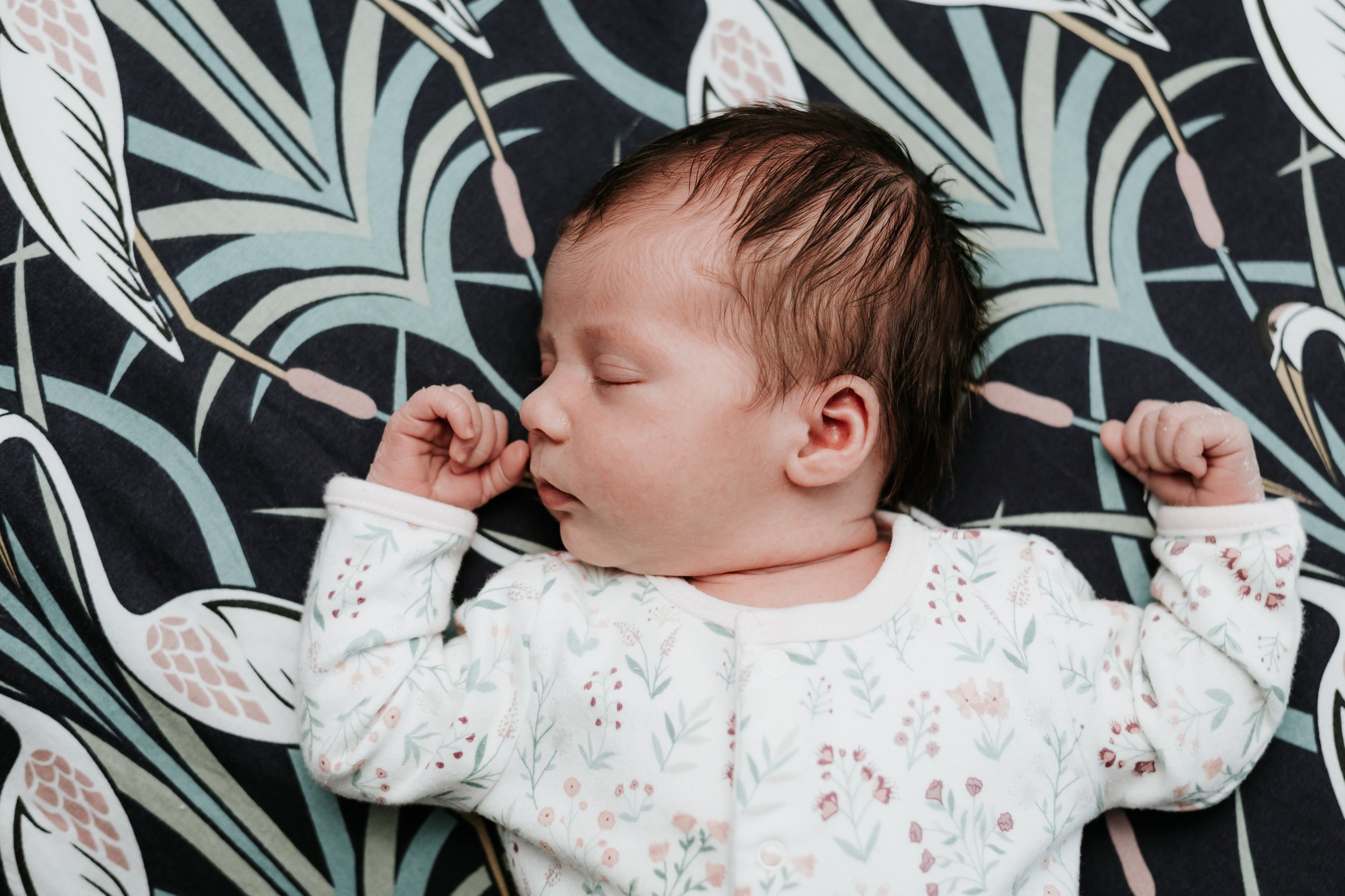 A sleeping baby with short, dark hair, dressed in a white onesie with a light floral pattern, lying on a dark fabric with a colorful leaf and plant pattern.