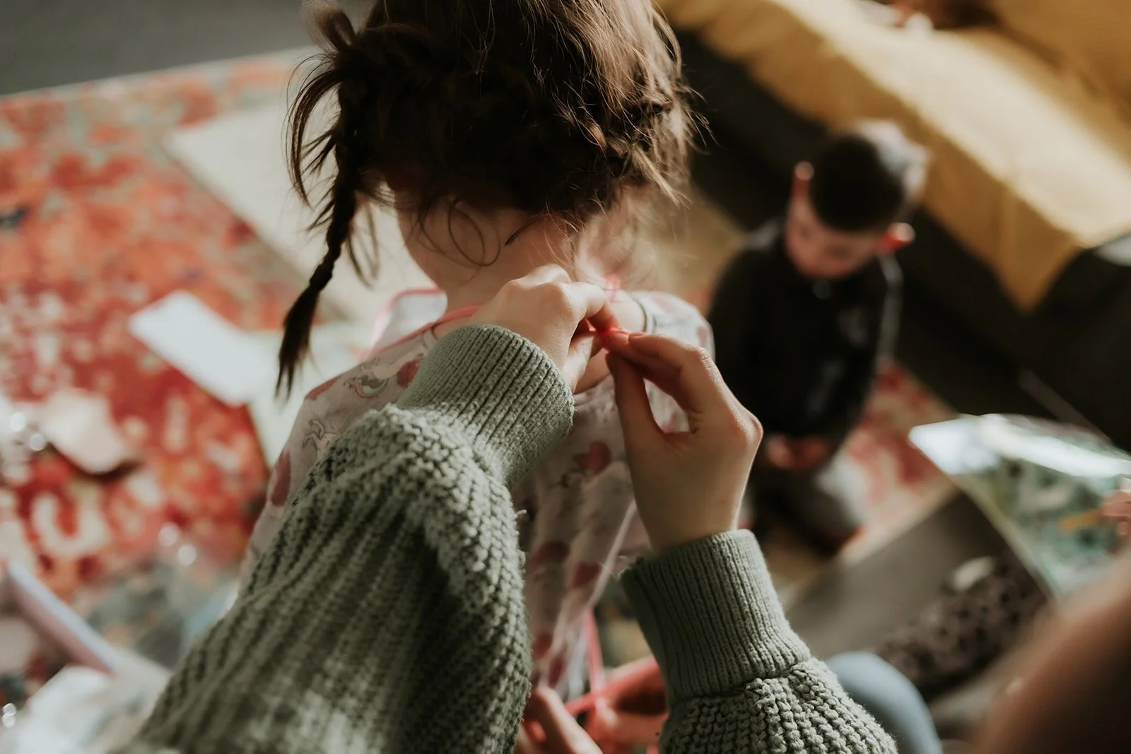 Person helping a young girl with a braided hairstyle, seated with a boy in the background in a cozy indoor setting.