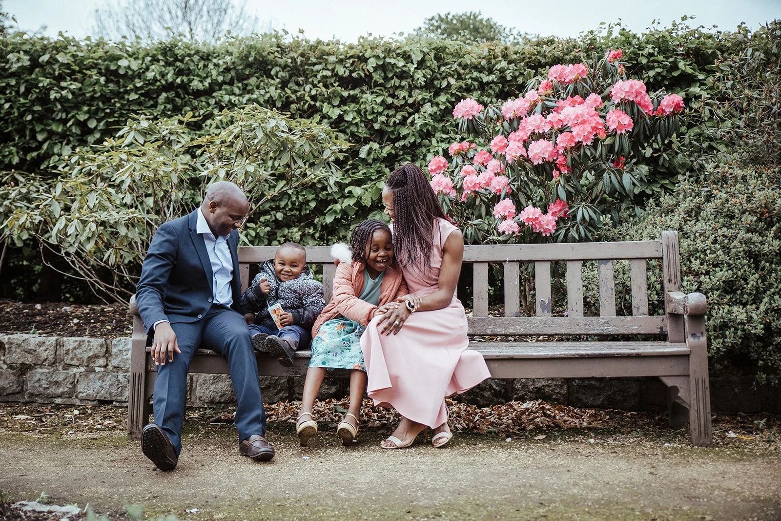 A family of four sitting on a park bench outdoors, surrounded by green bushes and pink flowers. The father is dressed in a suit, the mother in a pink dress, and their two children are sitting between them, smiling and laughing.