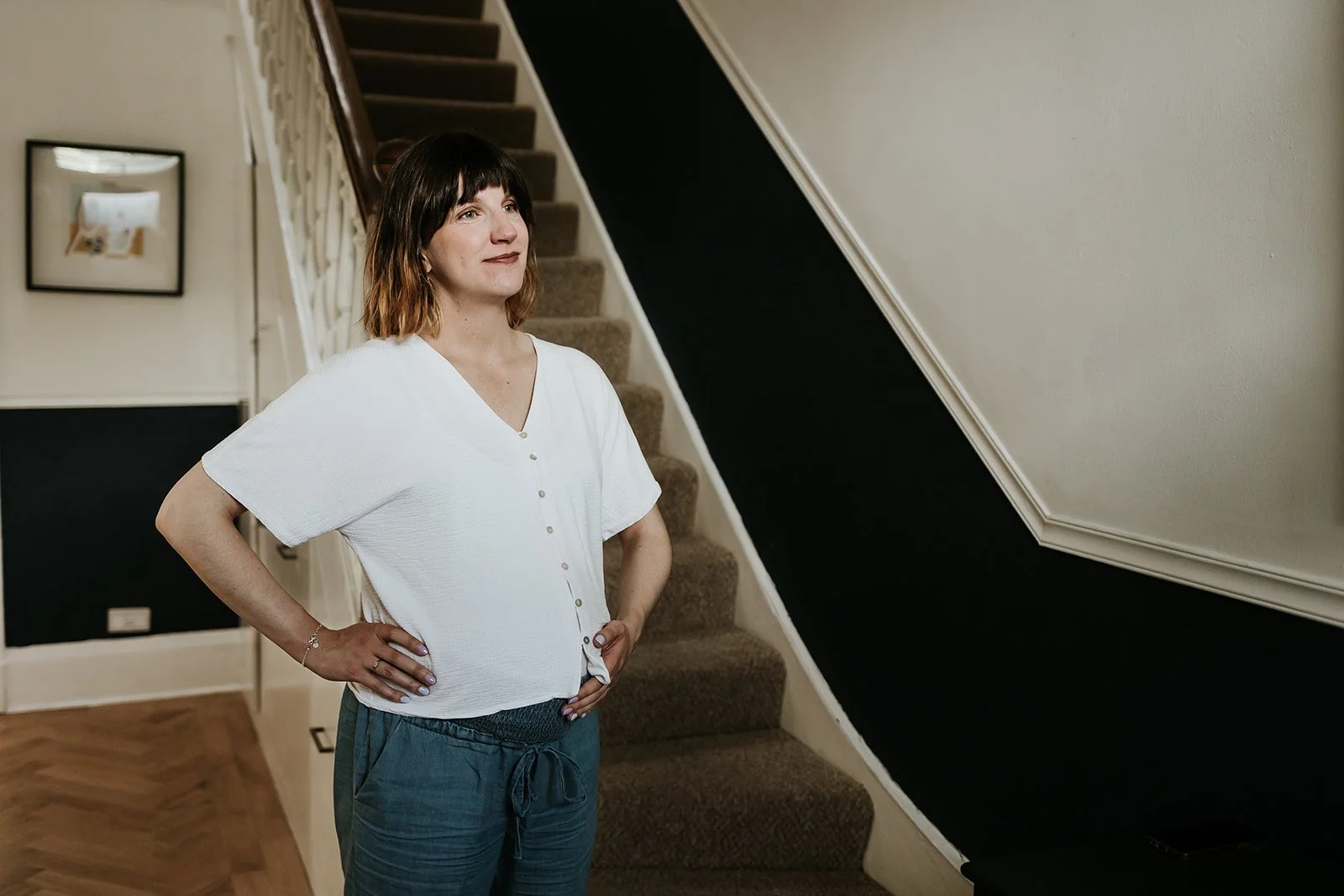 A woman with short dark hair, wearing a white button-down shirt and blue pants, stands with hands on hips in a home interior near a staircase with brown carpeted steps, a black-painted wall under the staircase, and framed picture on the wall.