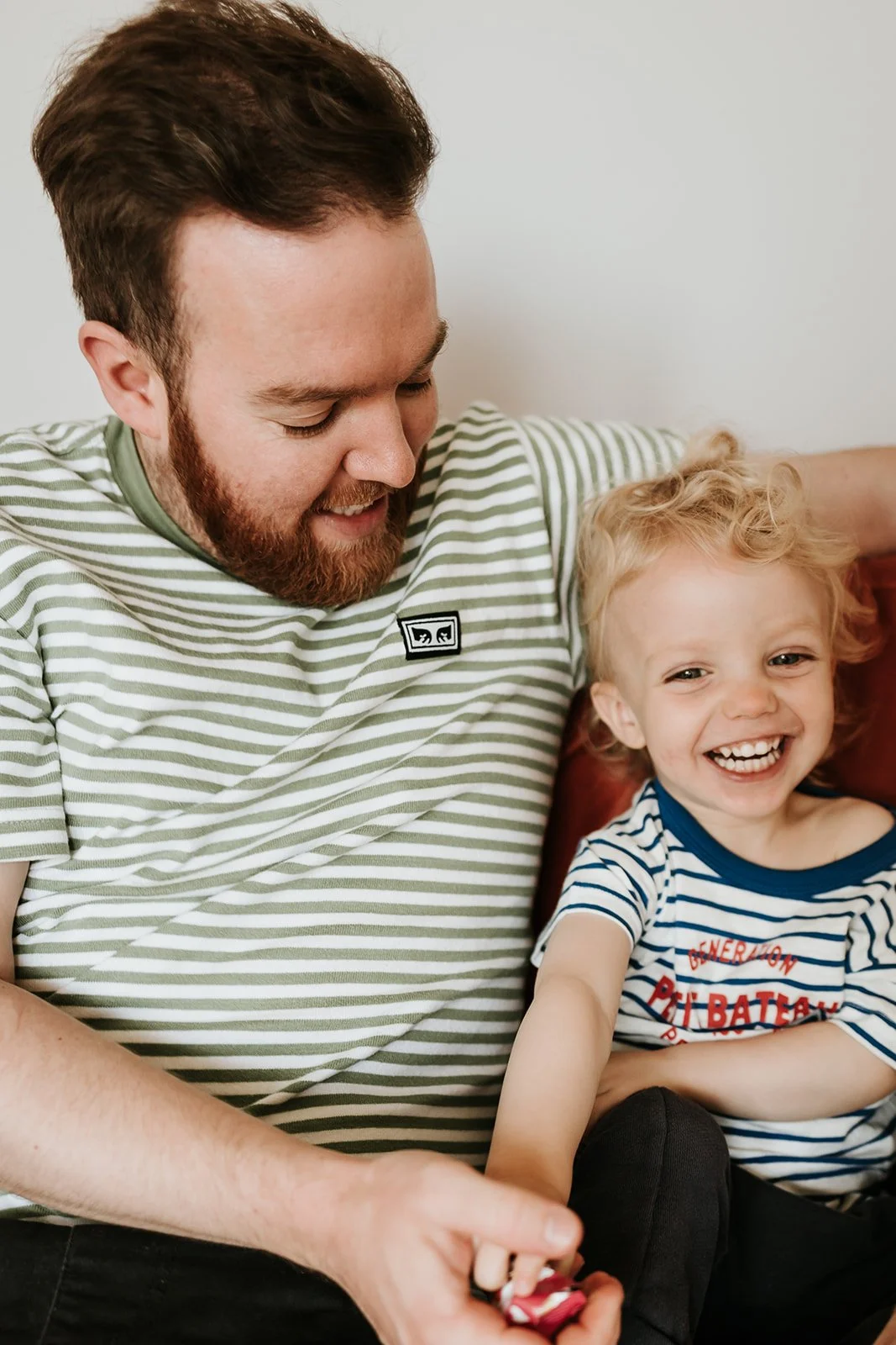 A man with a beard and a child with curly blonde hair sitting together, smiling and enjoying a happy moment.