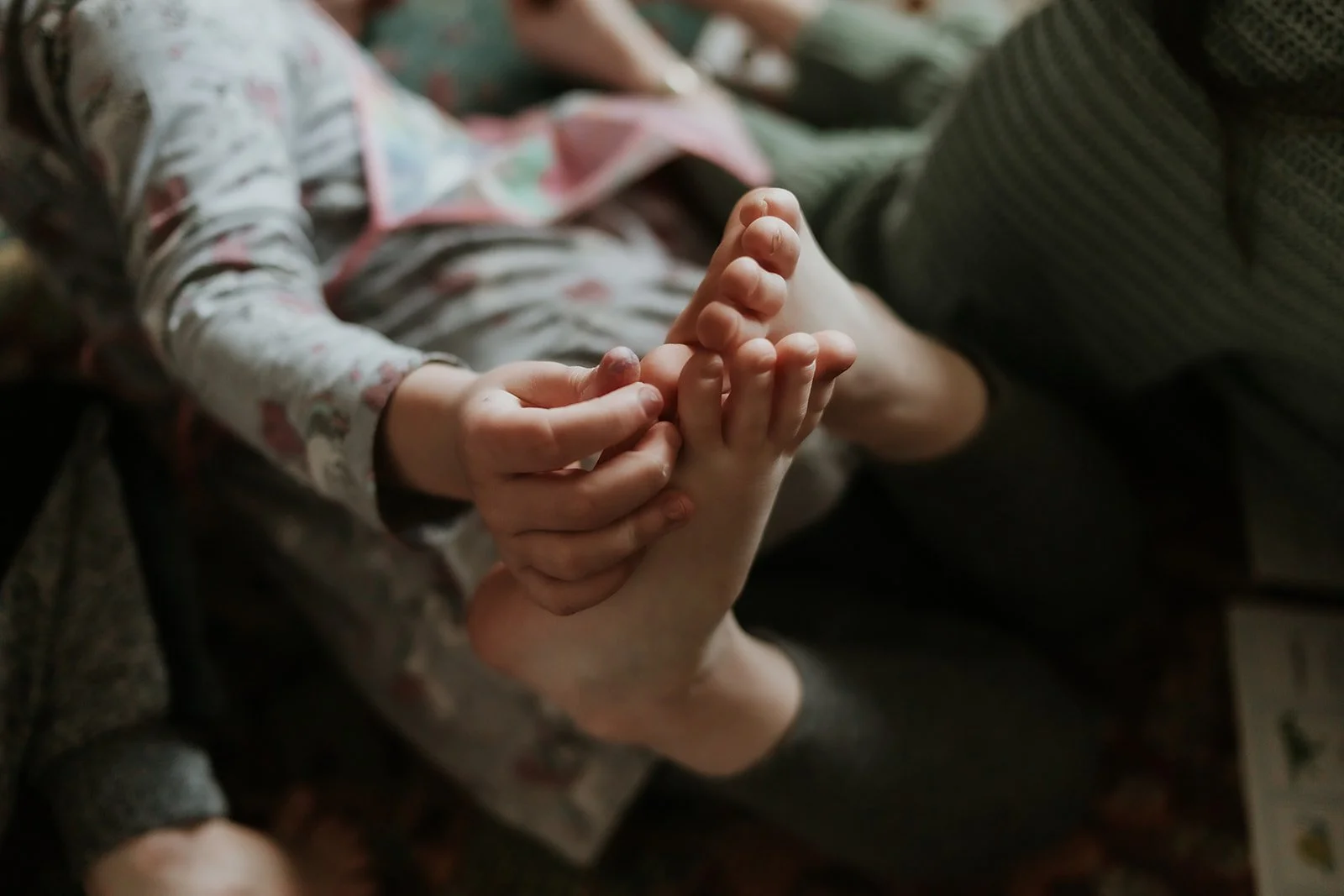 A child with a floral long sleeve shirt holding their own foot with both hands, while an adult, possibly a parent, gently touches the child's foot.