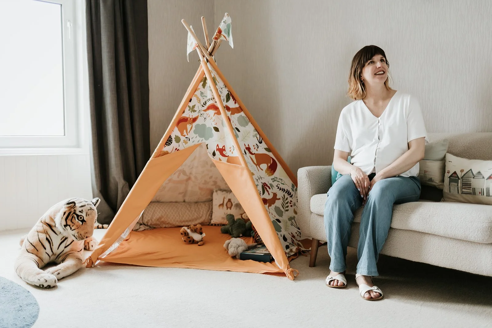 A woman sitting on a sofa in a living room with a children's play tent, stuffed animals, and a plush tiger toy on the floor.