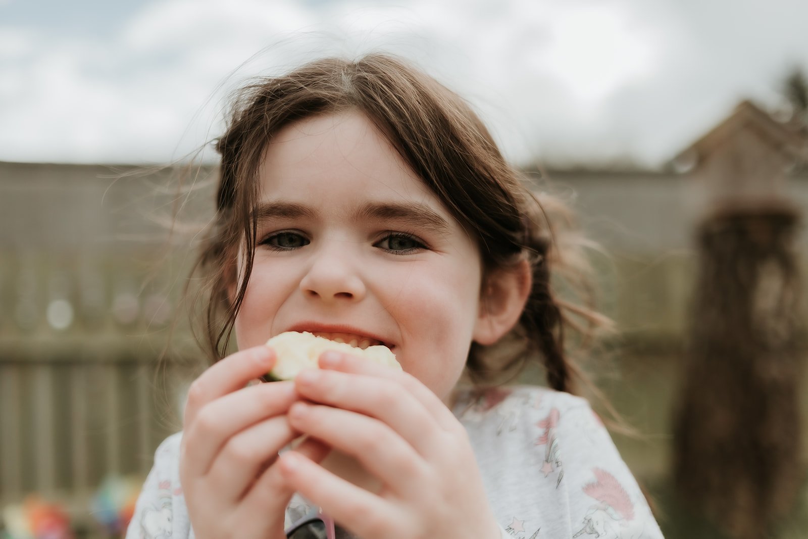 A young girl with brown hair and blue eyes is smiling as she about to eat a slice of apple outdoors, with a wooden fence and cloudy sky in the background.