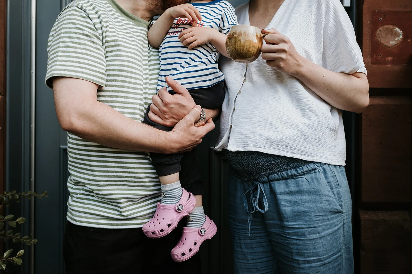 Three people, an adult man, a woman, and a small child, are standing close together. The man is holding the child, who is wearing pink Crocs and striped socks. The woman is holding a wooden mug. They are standing in front of a dark wooden wall.