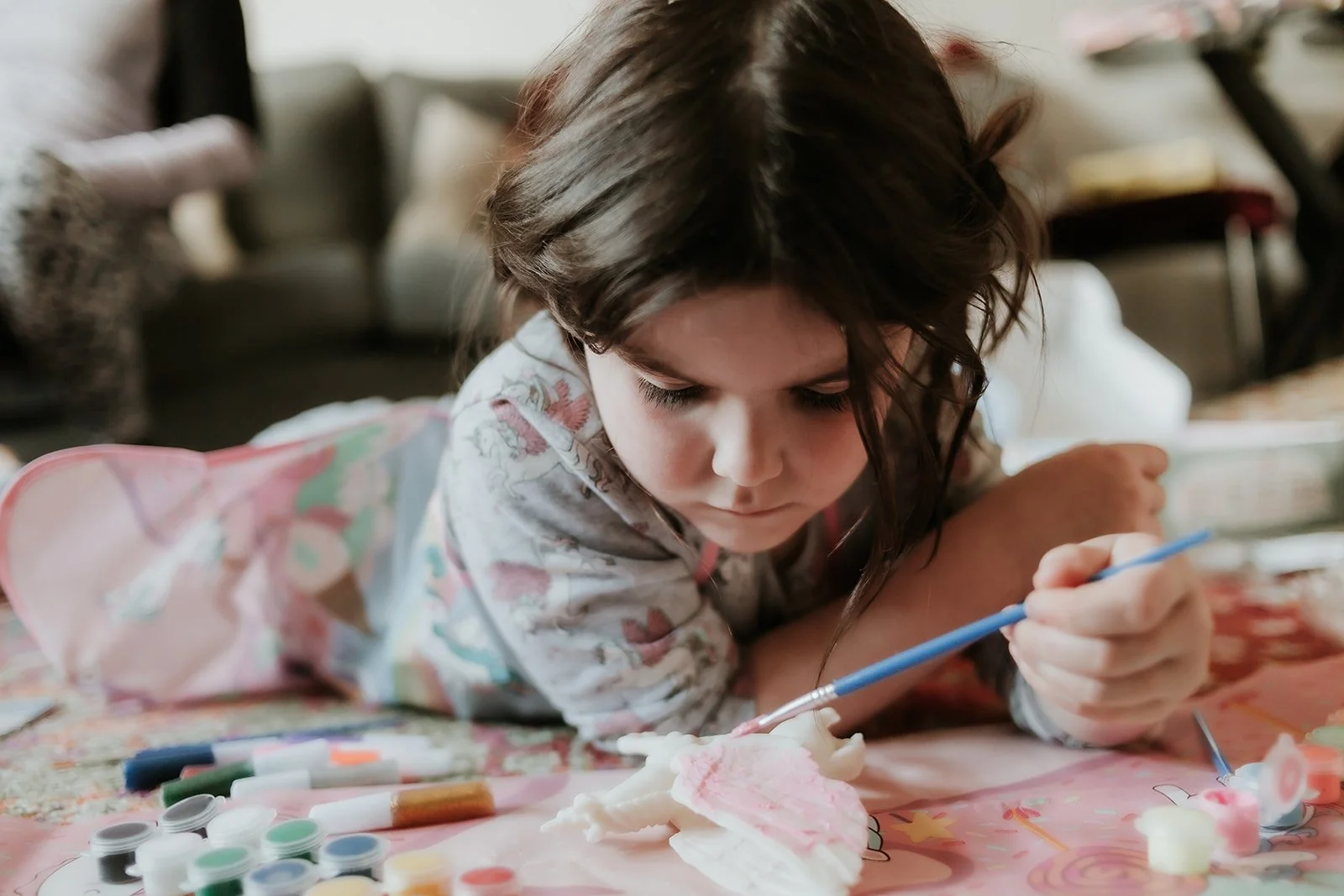 Young girl lying on a colorful table, painting a ceramic figurine with a small blue brush, surrounded by paint bottles and art supplies in a cozy, well-lit room.
