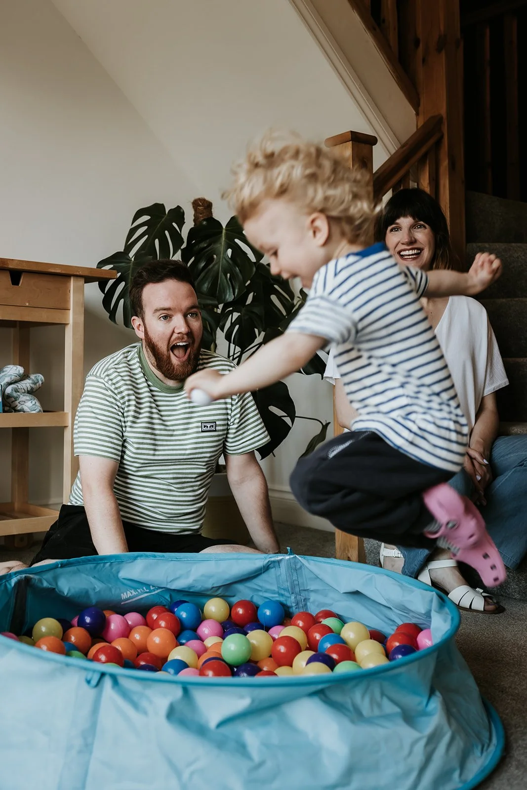 A jumping child with blonde hair in striped shirt and pink Crocs playing with colorful balls in a blue ball pit, while two adults, a man and a woman, watch and smile in a cozy home setting.