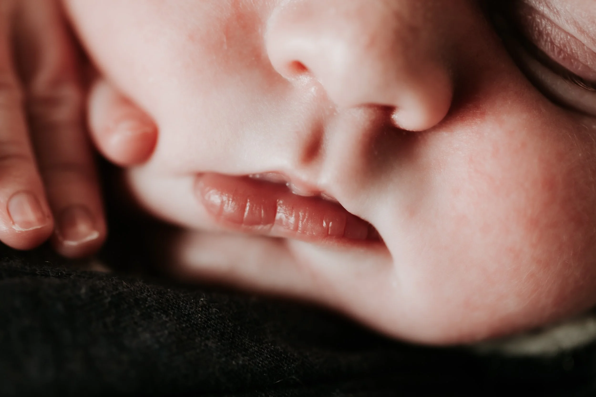 Close-up of a baby's face with eyes closed, lying on a black surface, being gently cradled or touched by an adult's hand.