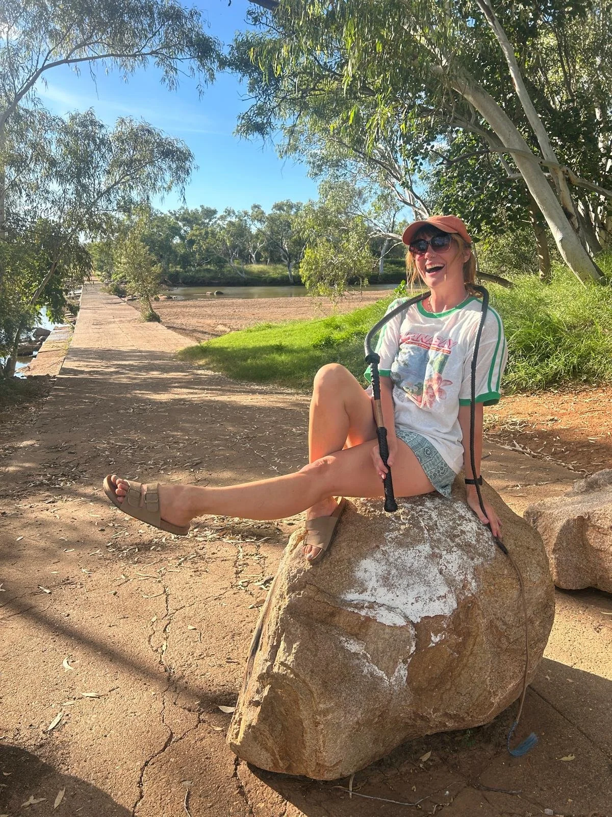 A woman sitting on a large rock near a riverbank, smiling and enjoying a sunny day in a park. She is wearing sunglasses, a hat, a casual T-shirt, shorts, and sandals, with a walking stick resting on her lap. The background features trees, a pathway, and a body of water.