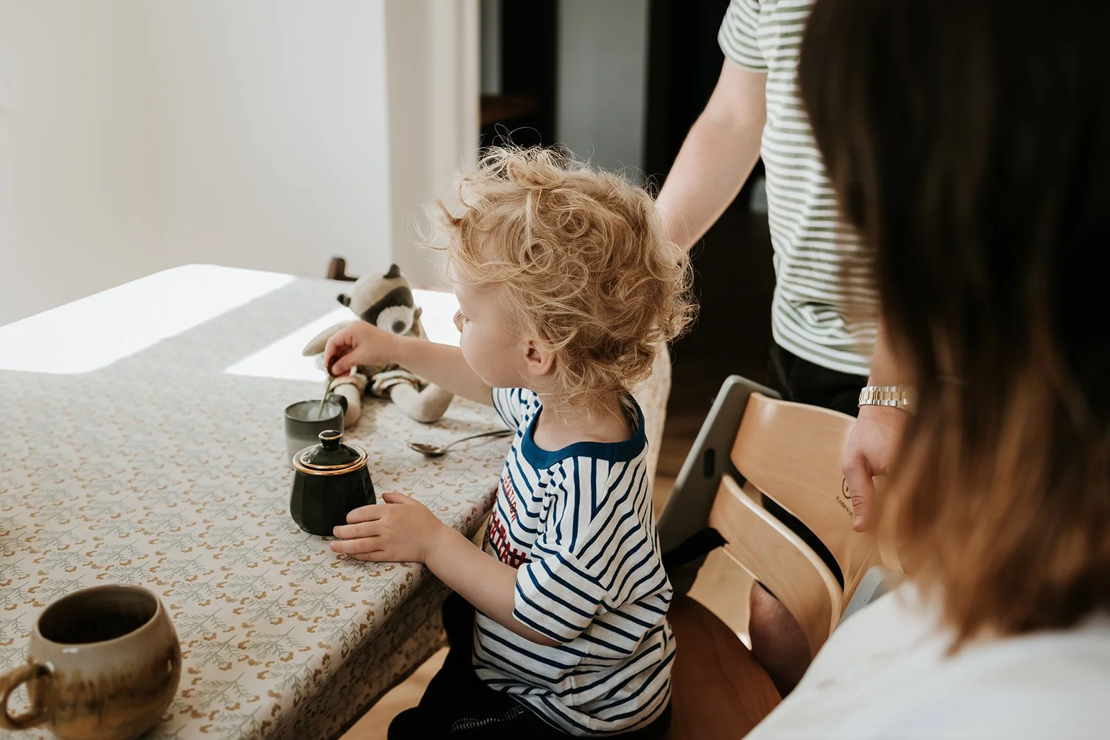 A young boy with curly blond hair wearing a striped shirt sitting at a dining table with a tea set and a stuffed animal, while an adult woman watches nearby.
