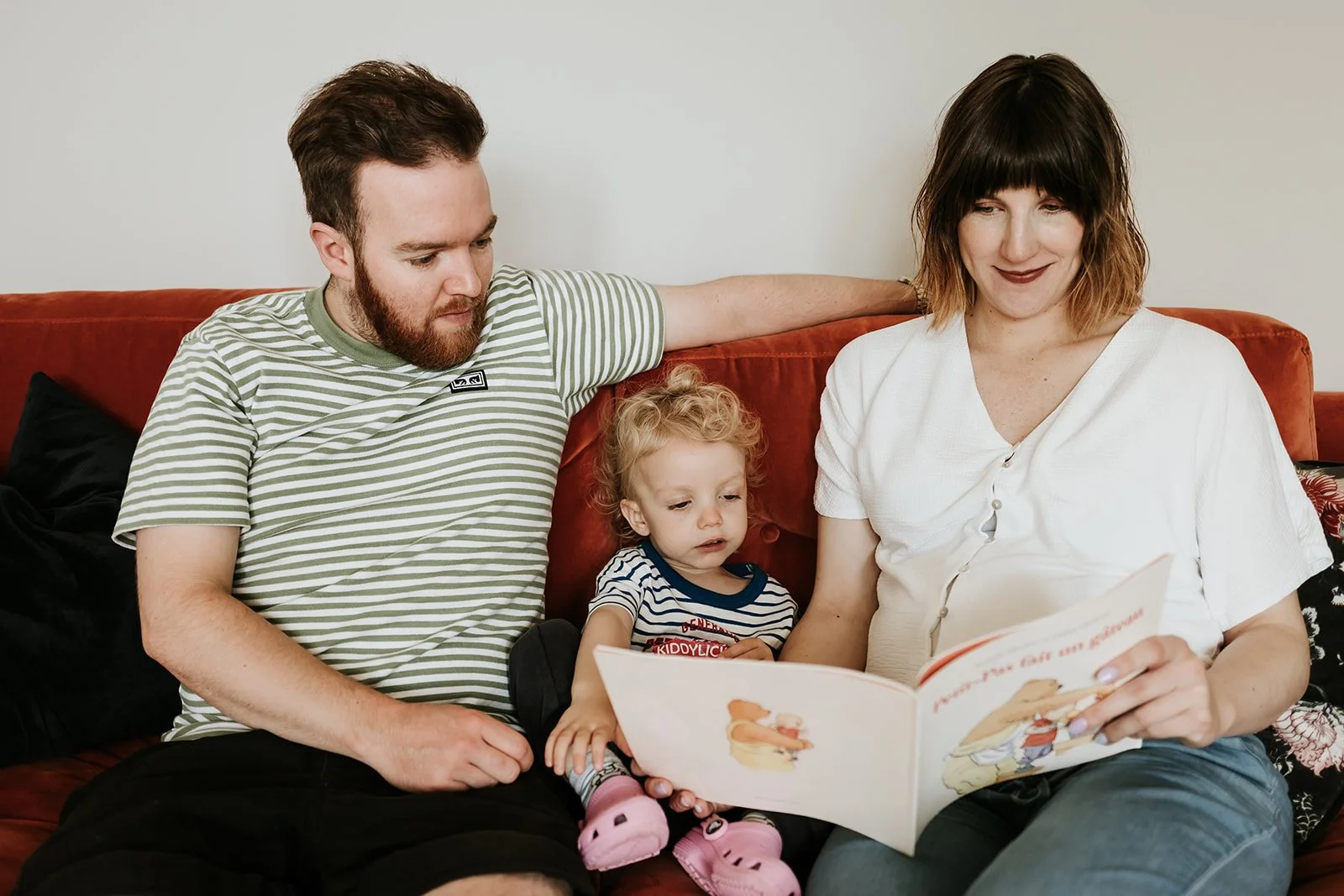 A family sitting on a couch, with a man, woman, and young child, reading a children's book together.