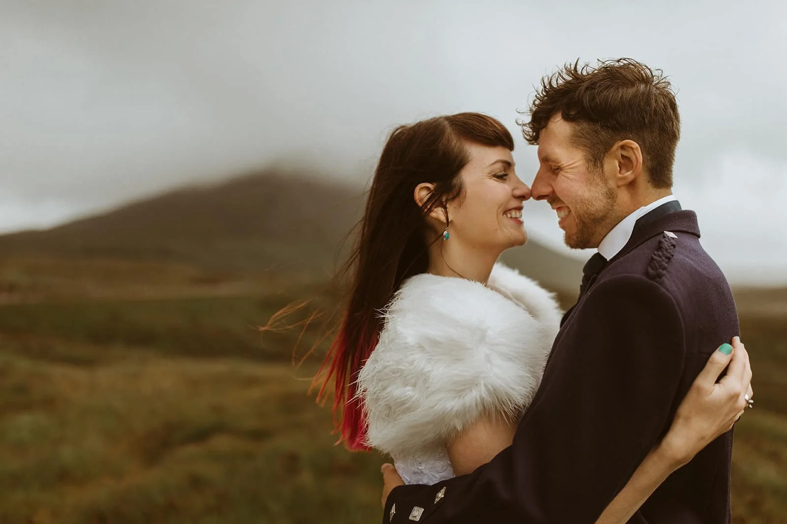 A happy couple is embracing outdoors in a grassy, hilly landscape with a cloudy sky, smiling and touching noses.