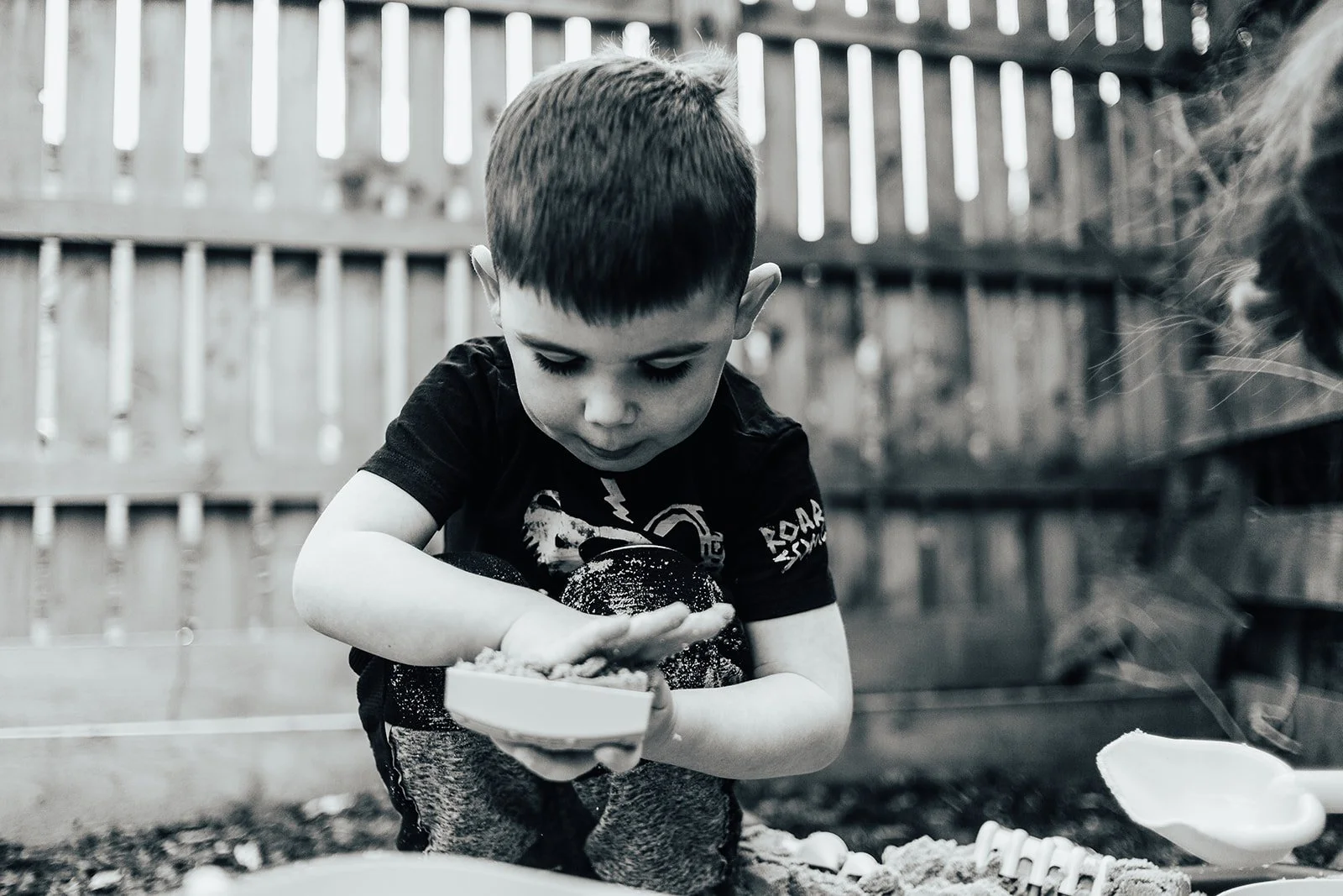 A young boy playing with sand outdoors, sitting on the ground with a bowl in his hands, surrounded by a wooden fence.