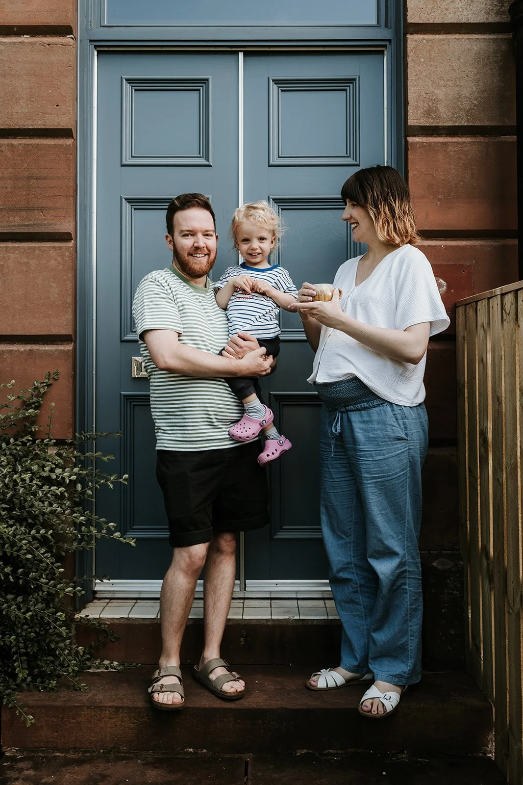A family of three standing outside a blue door. The father is holding a young girl, and the mother is holding a bowl and smiling at her family.