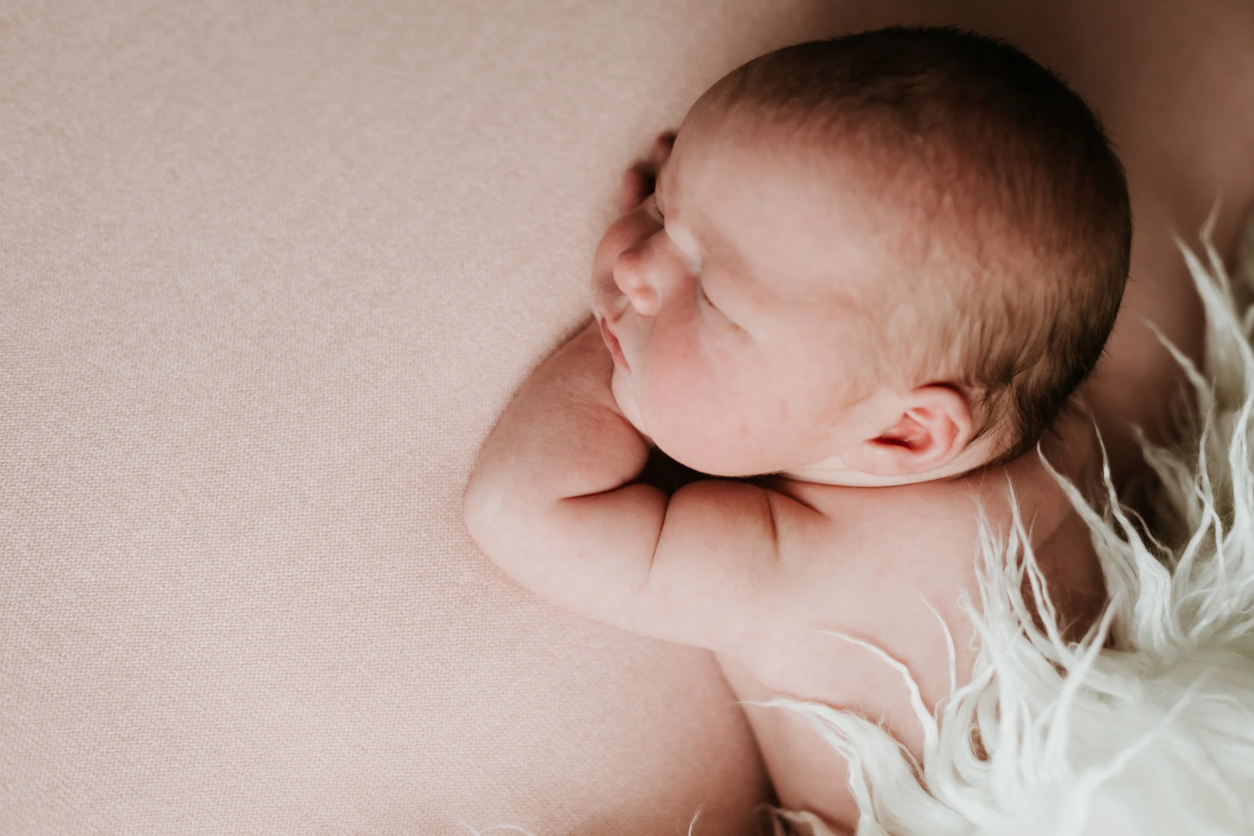 Close-up of a sleeping newborn baby with a fade hairstyle, resting on a soft pink surface, wearing white feathered clothing.