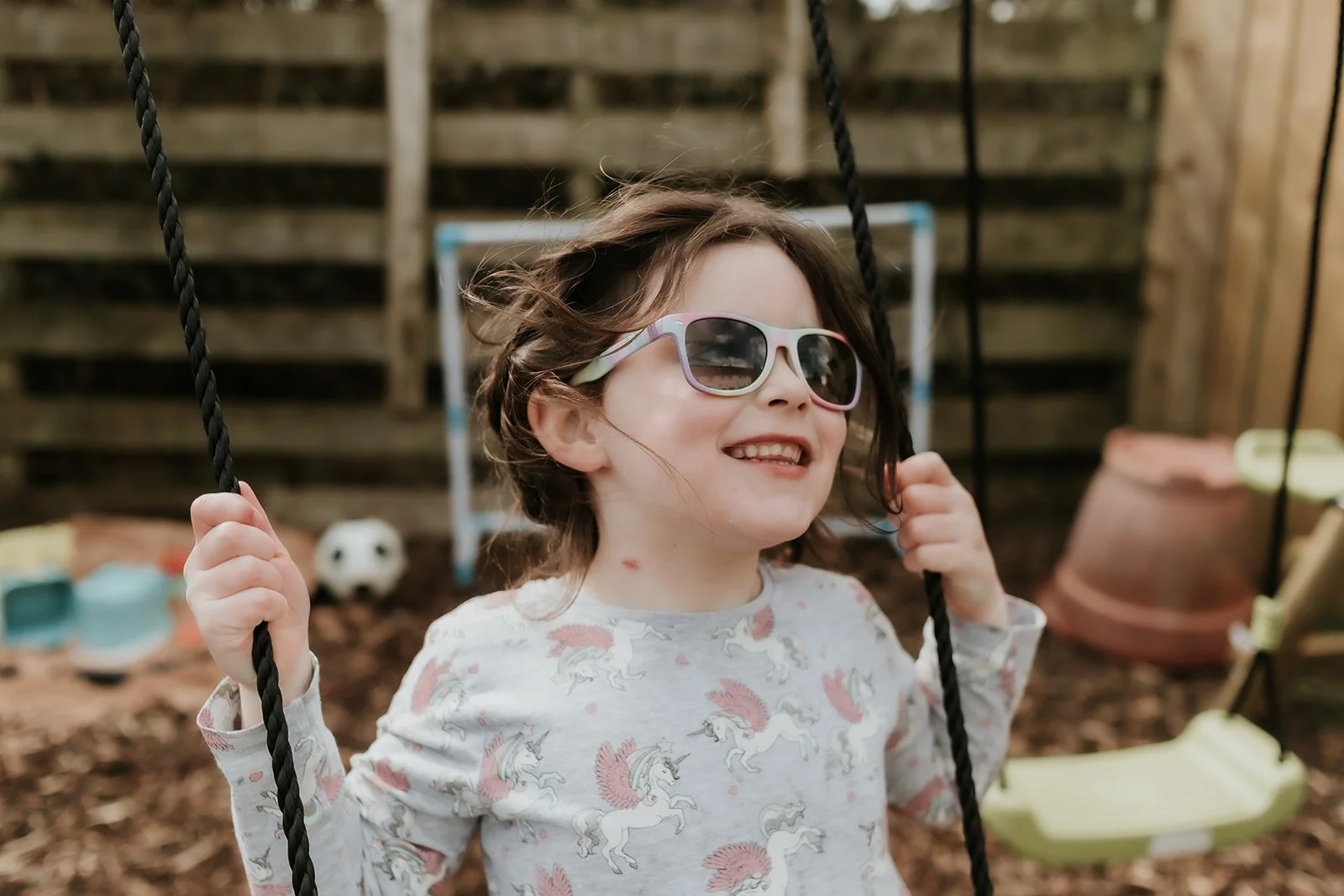A young girl with brown hair, wearing sunglasses and a unicorn-printed shirt, smiling on a swing set in a backyard.