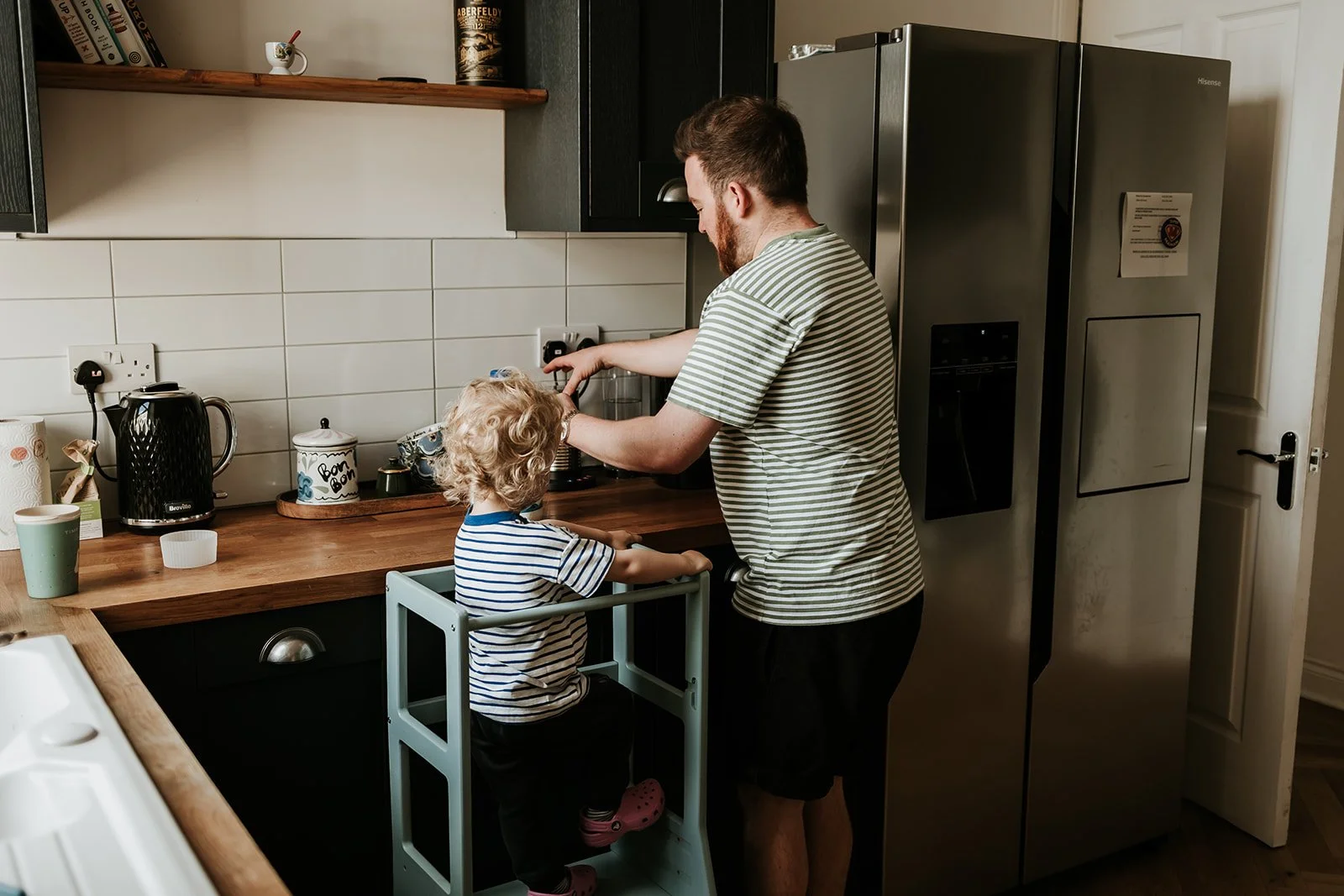 A man and a young girl in a kitchen, with the man helping the girl reach the countertop, which is at a standing height for him, while she stands in a toddler safety stand. The man is wearing a striped t-shirt and black shorts, and the girl is wearing