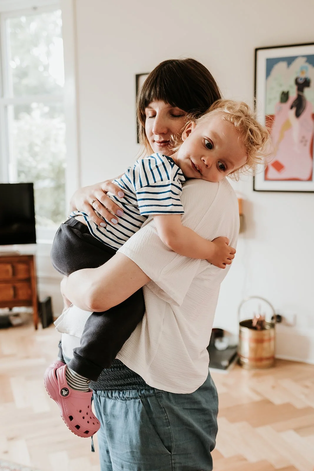 A woman is hugging a young child in a cozy, well-lit living room. The woman has short dark hair and is wearing a white t-shirt, while the child has blonde curly hair, is wearing a striped shirt and pink crocs. The background includes a window, artwork on the wall, and a wooden table.