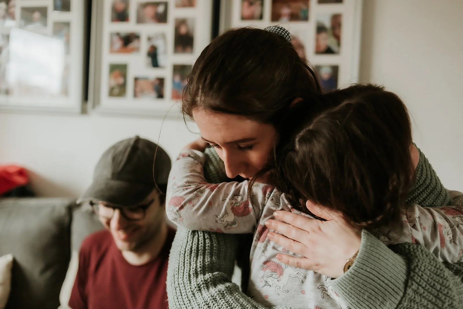 A woman with dark hair hugging a child with brown hair in a cozy living room, with a man wearing glasses and a cap smiling in the background.