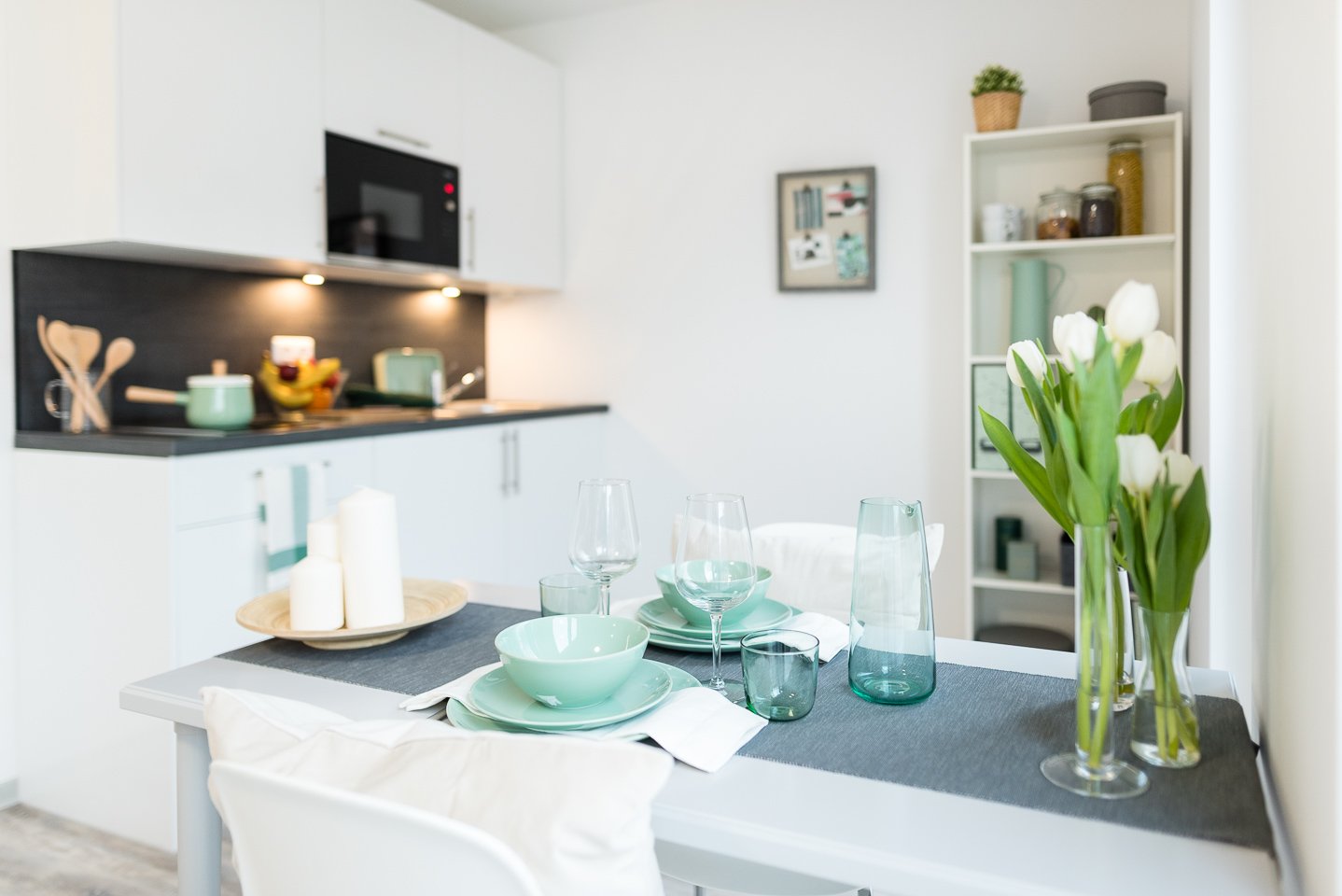Moderne Küche mit Esstisch, gedeckt mit türkisem Geschirr und Gläsern. Weiße Tulpen im Hintergrund. Weiße Möbel mit schwarzer Arbeitsplatte. Dekorative Details wie Kerzen und Pflanzen.